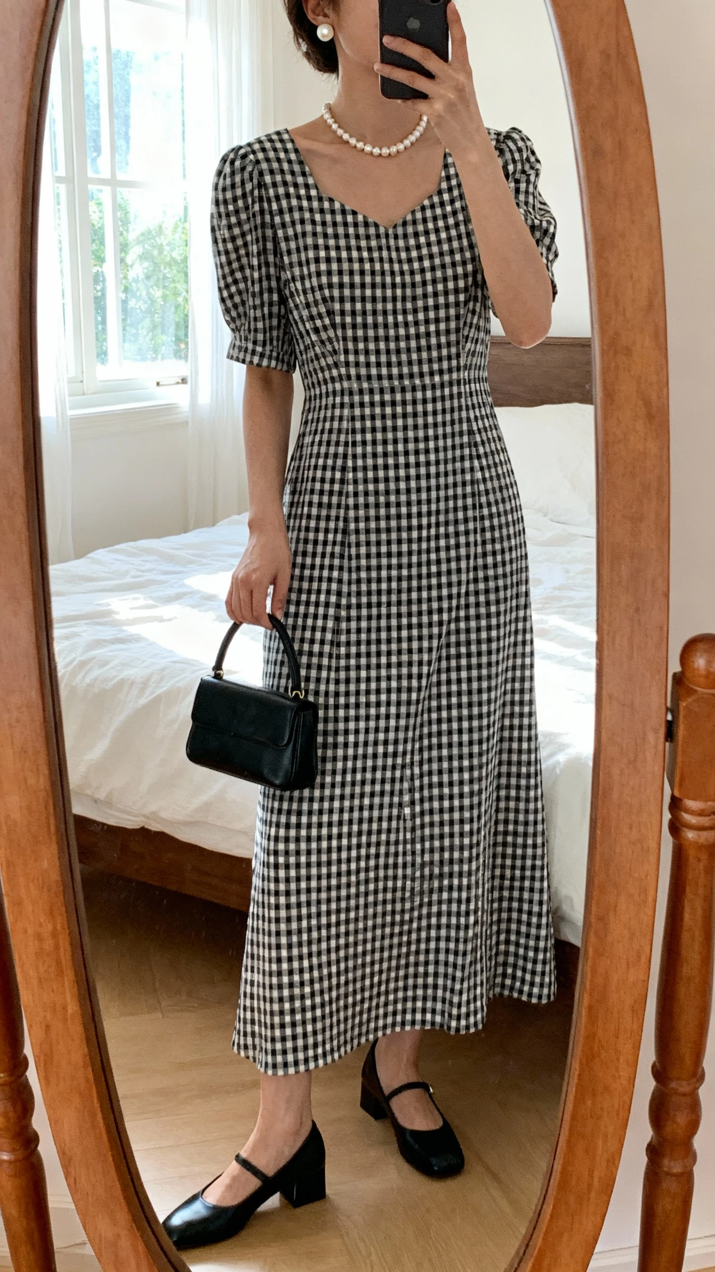 iPhone mirror selfie of a woman wearing a classic black-and-white gingham full-length dress with a modest neckline and sleeves, single-strand pearl necklace, pearl stud earrings, low block-heel black flats, and a small black top-handle bag, face not visible, cropped at neck, bedroom mirror, natural window light, iPhone photo quality.