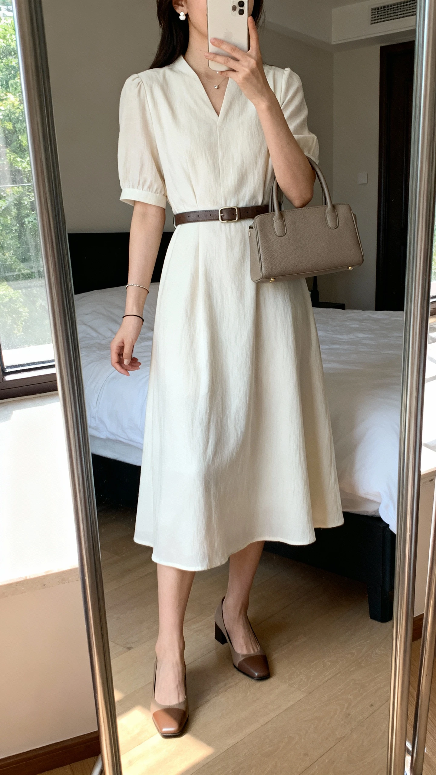 iPhone mirror selfie of a woman wearing a classic midi dress with a belted waist, low block heels, structured top-handle bag, delicate stud earrings, and a thin bracelet, face not visible, cropped at neck, bedroom mirror with natural window light, iPhone photo quality.