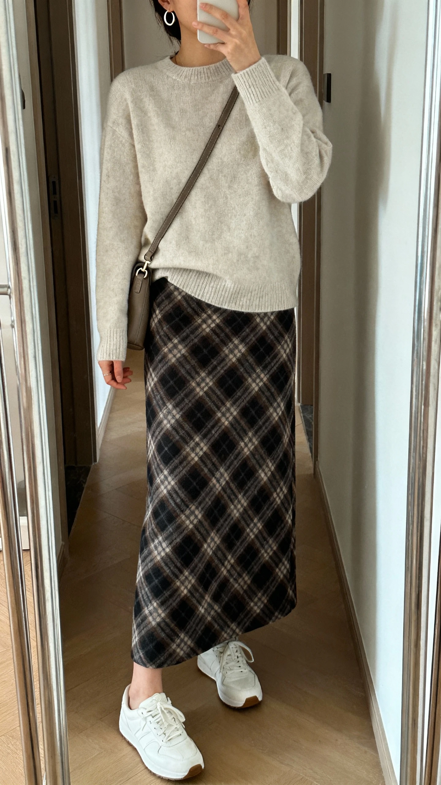 iPhone mirror selfie of a woman wearing a classic plaid long skirt with a soft crewneck sweater, clean white sneakers, crossbody bag, and dainty hoop earrings, face not visible, cropped at shoulders, hallway full-length mirror, natural daylight, iPhone photo quality.