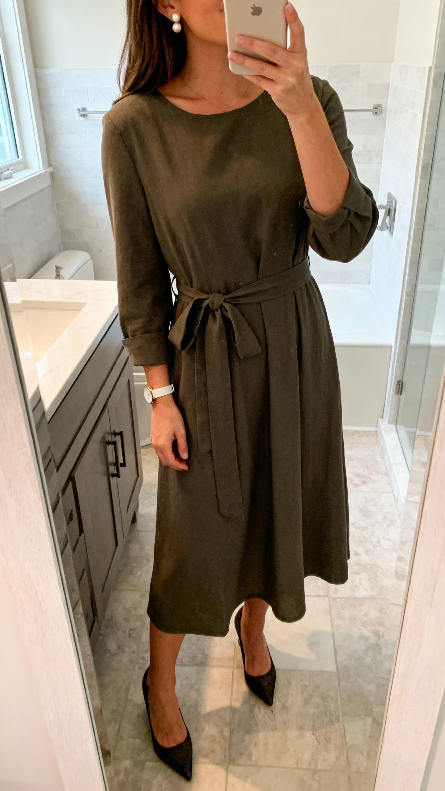 iPhone mirror selfie of a woman wearing a classic sleeved midi dress with a waist tie, kitten heels, pearl stud earrings, and a minimal wristwatch, face not visible, cropped at shoulders, bathroom mirror, soft natural daylight, iPhone photo quality.