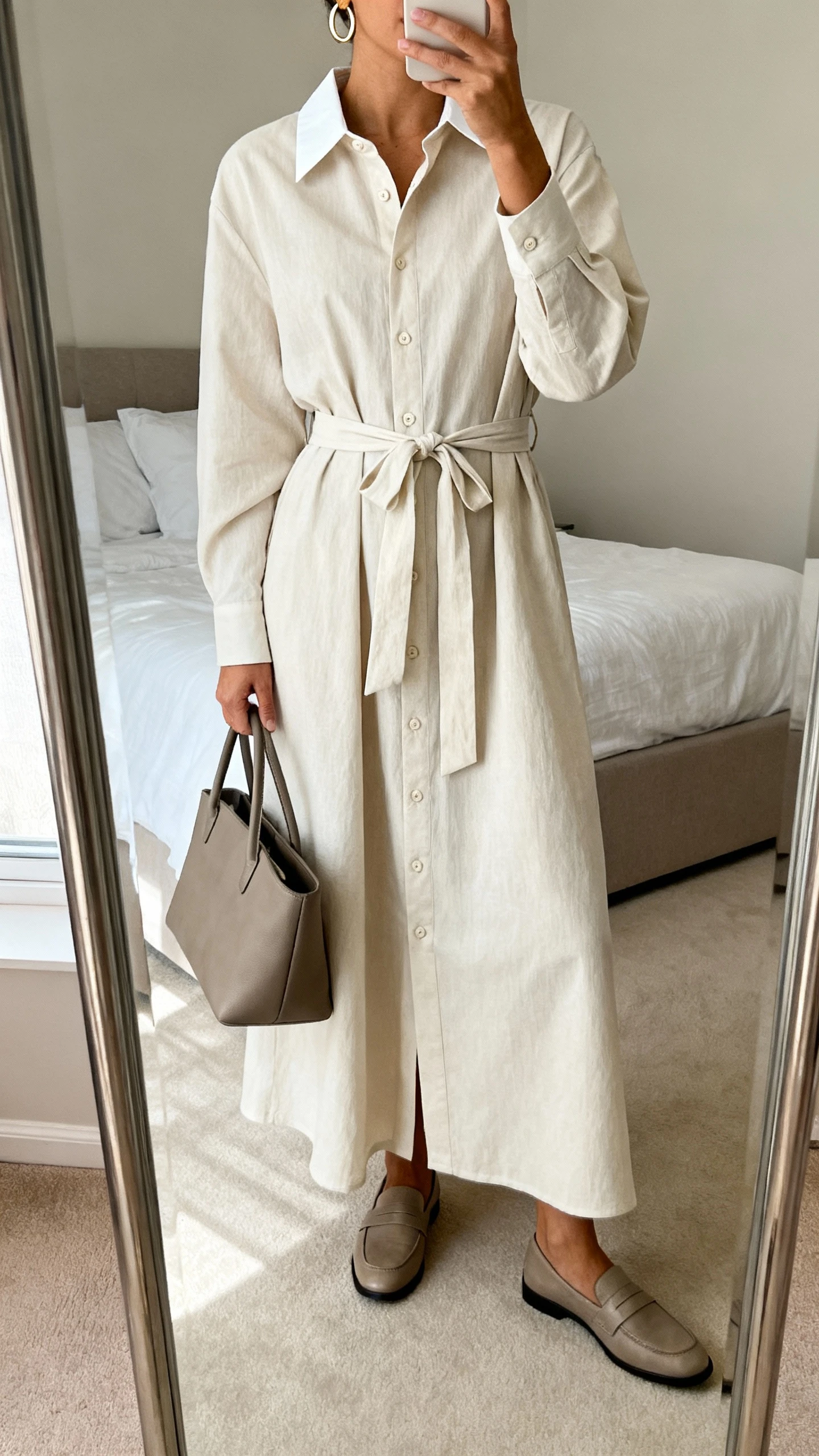 iPhone mirror selfie of a woman wearing a classic shirt-dress maxi with a crisp collar, button front, self-tie belt, simple hoop earrings, structured tote, and loafers, face not visible, cropped at neck, bedroom mirror, soft natural light, iPhone photo quality