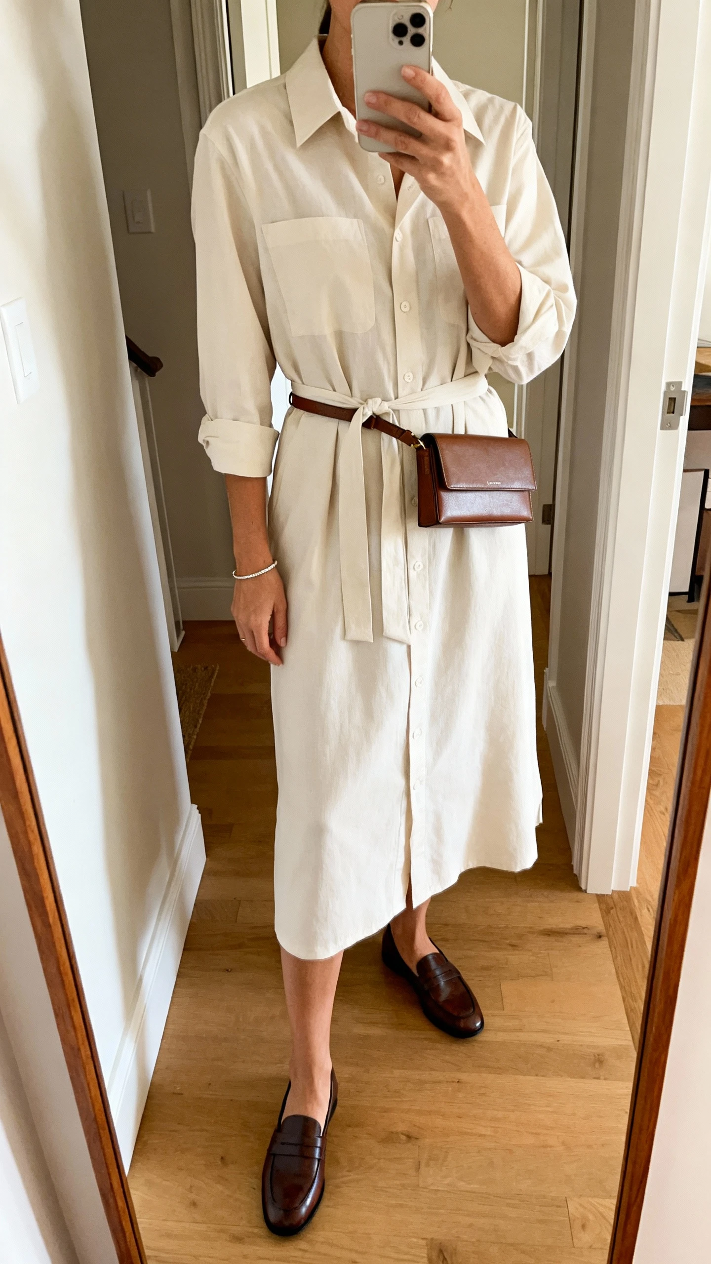iPhone mirror selfie of a woman wearing a classic shirt dress with a tie belt, midi length, loafers, slim leather belt bag, simple bracelet, face not visible, cropped at neck, entryway full-length mirror, warm indoor lighting, iPhone photo quality