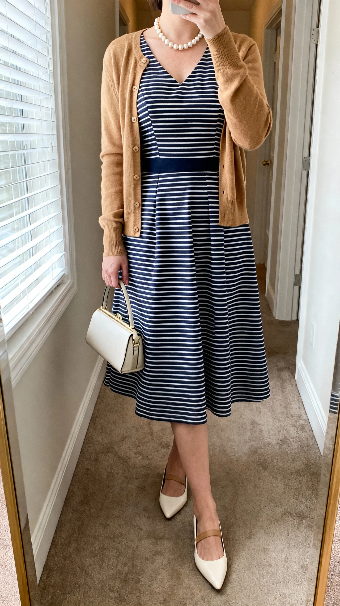 iPhone mirror selfie of a woman wearing a classic striped Sunday-best dress (navy stripes on white, tailored bodice, modest midi A-line), with a camel cardigan, pearl necklace, pointed flats, and a small top-handle bag; face not visible, cropped at neck, hallway full-length mirror, natural window light, iPhone photo quality.
