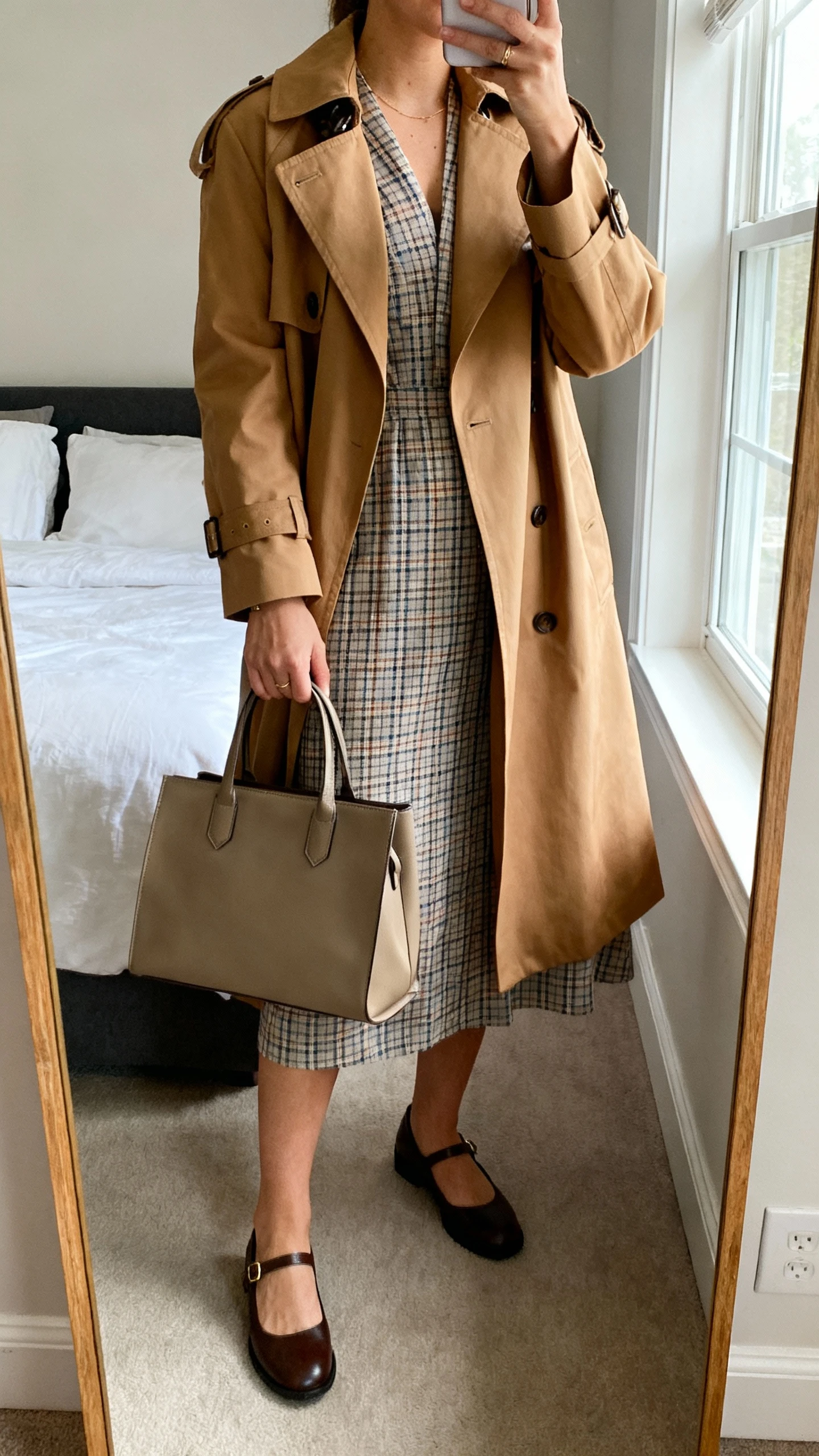 iPhone mirror selfie of a woman wearing a classic plaid dress paired with Mary Janes and a camel trench coat, structured tote bag in hand; face not visible, cropped at neck, bedroom mirror, natural window light, iPhone photo quality