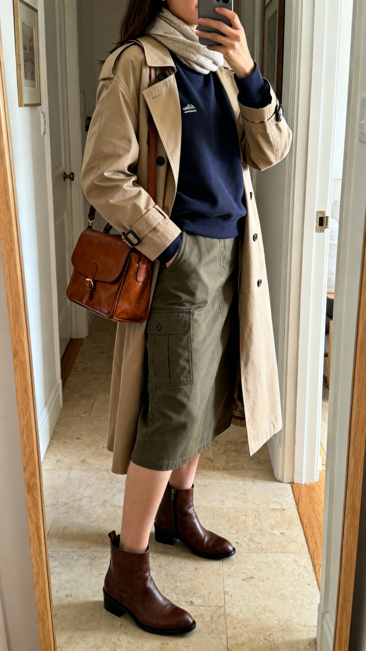 iPhone mirror selfie of a woman wearing a classic crewneck with a utility skirt, trench coat layered on top, ankle boots, leather satchel, and a simple scarf; face not visible, side angle, entryway full-length mirror, indoor natural light, iPhone photo quality.