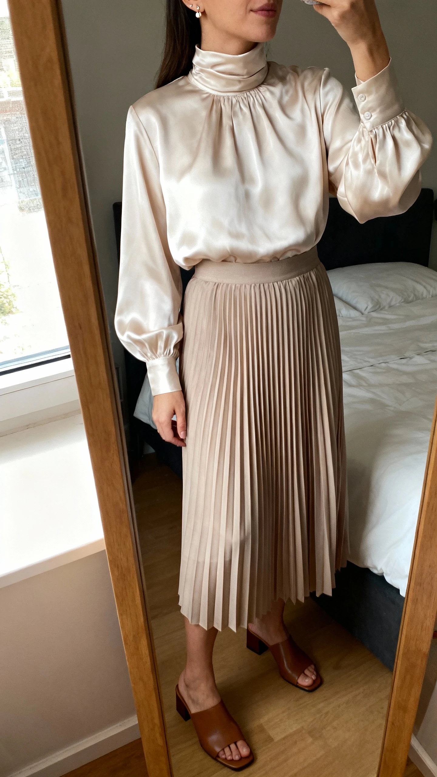 iPhone mirror selfie of a woman wearing a cloud-soft high-neck satin blouse tucked into a pleated midi skirt with delicate stud earrings and low block-heel mules, face not visible, cropped at neck, bedroom mirror, natural window light, iPhone photo quality.