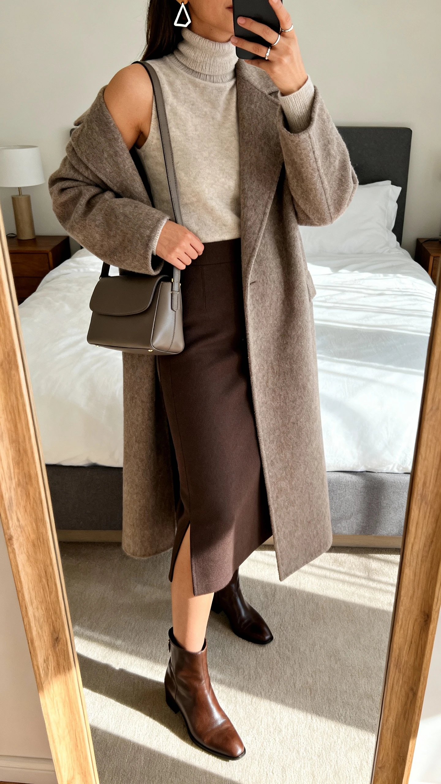 iPhone mirror selfie of a woman wearing a column midi skirt with a back vent, a lightweight turtleneck, a longline wool coat, leather ankle boots, a sleek shoulder bag, geometric earrings, and a minimal ring, face not visible, cropped at shoulders, bedroom mirror, soft afternoon light, iPhone photo quality.