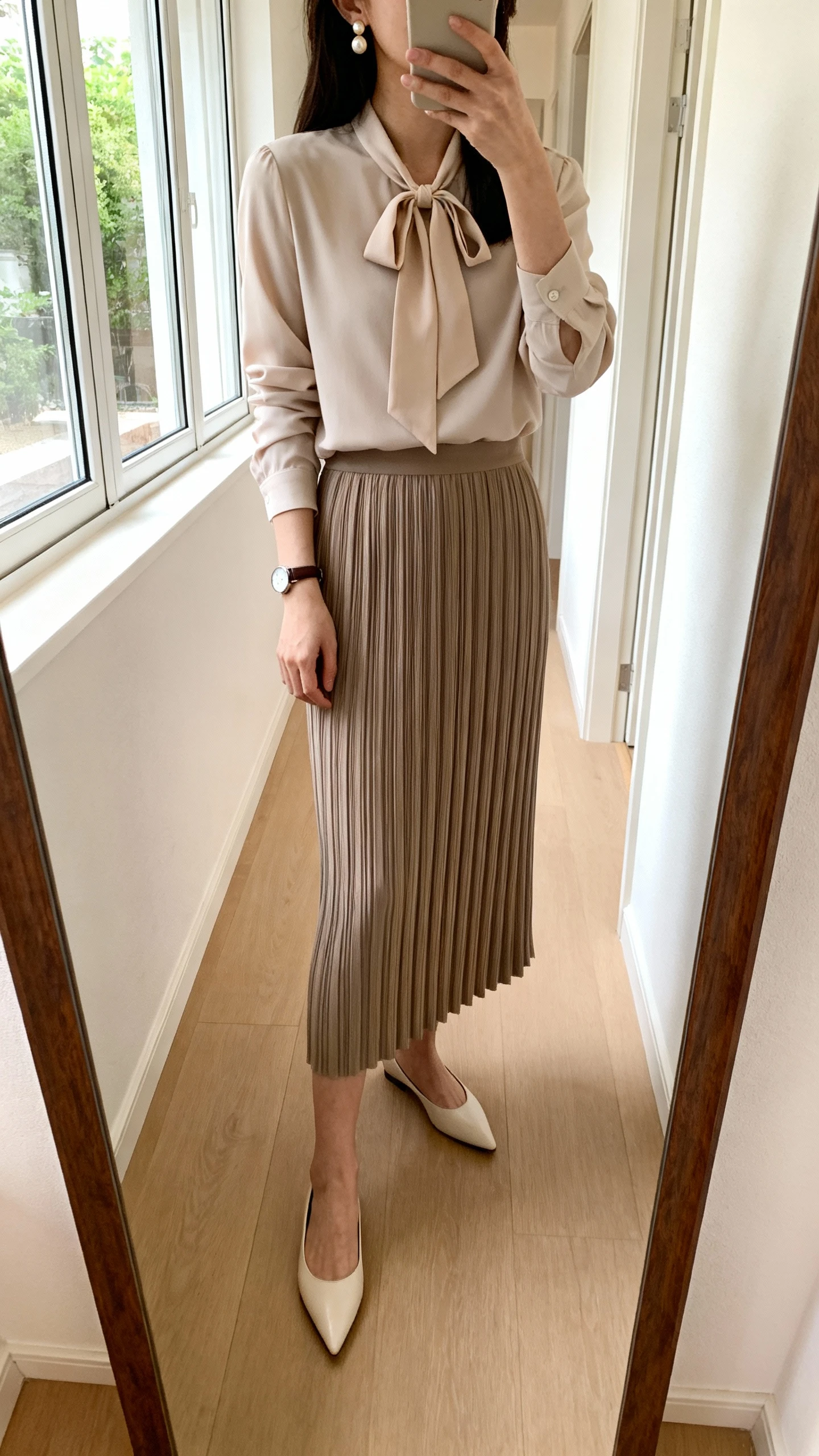iPhone mirror selfie of a woman wearing a column skirt and a long-sleeve blouse with a gentle bow tie at the neck, pearl earrings, pointed-toe flats, and a simple wristwatch, face not visible, cropped at shoulders, hallway mirror, soft window light, iPhone photo quality.
