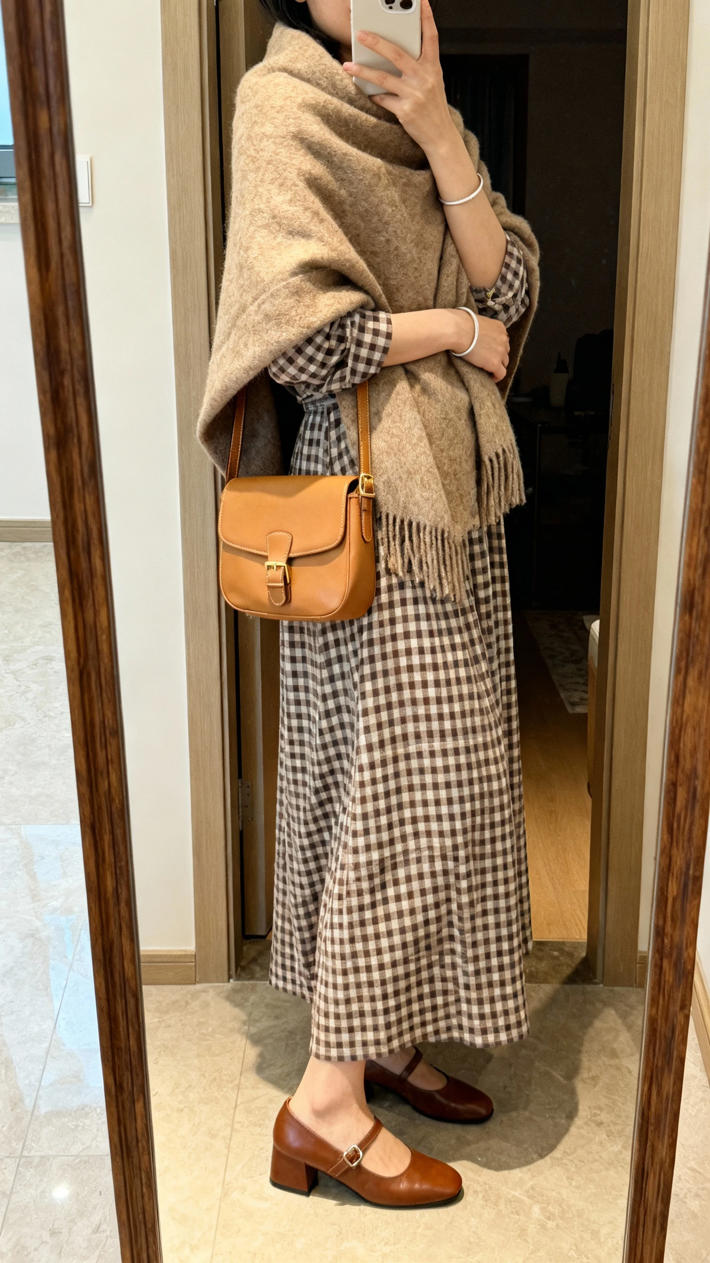 iPhone mirror selfie of a woman wearing a country-chic gingham maxi dress wrapped in a cozy wool shawl, camel crossbody satchel, brown low-heel Mary Janes, and a simple bracelet, face not visible, side angle, entryway mirror, warm indoor lighting, iPhone photo quality.