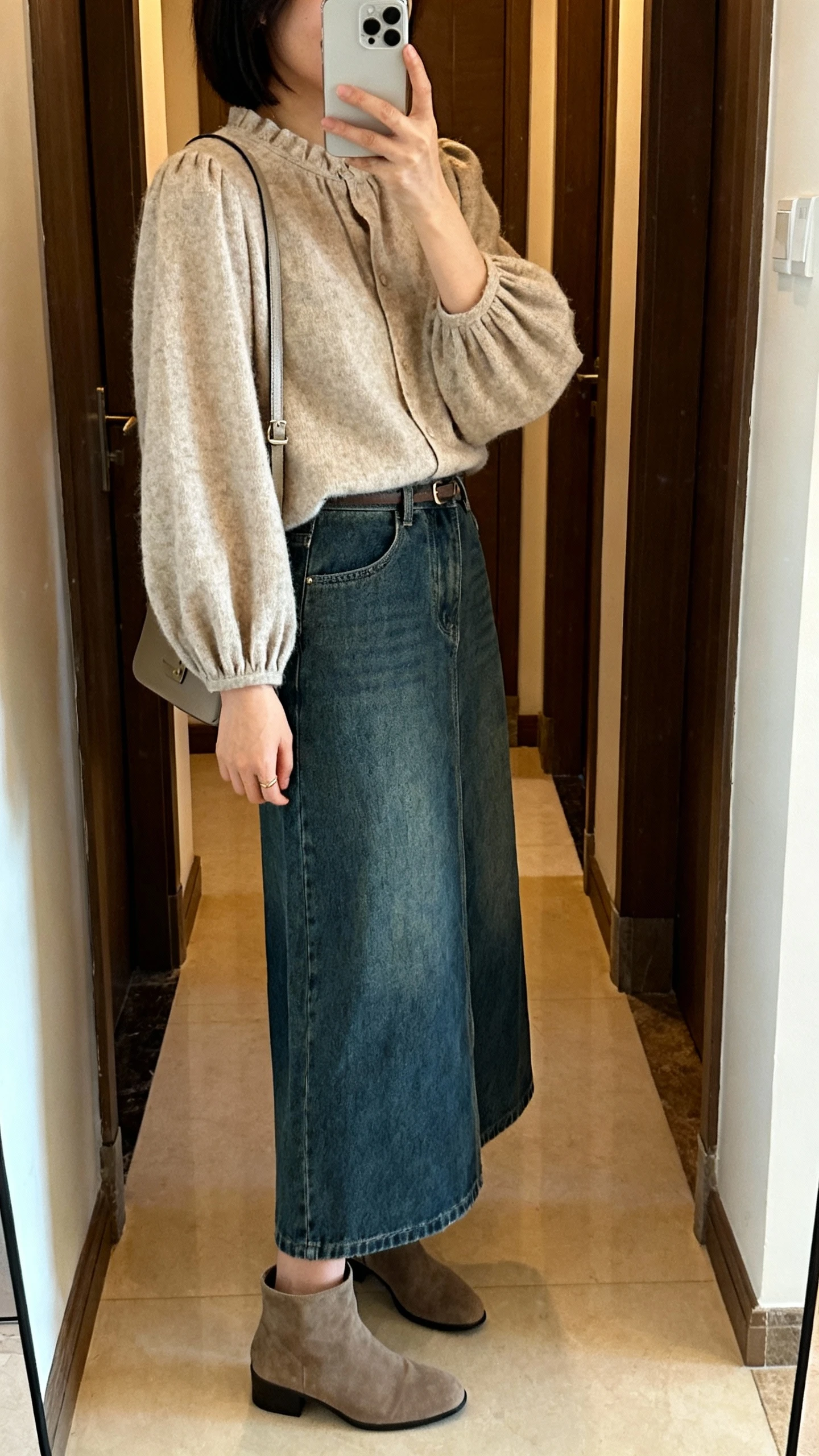 iPhone mirror selfie of a woman wearing a cozy bishop-sleeve blouse paired with a denim maxi skirt, ankle boots and a simple crossbody bag, face not visible, side angle, entryway mirror, indoor warm lighting, iPhone photo quality.