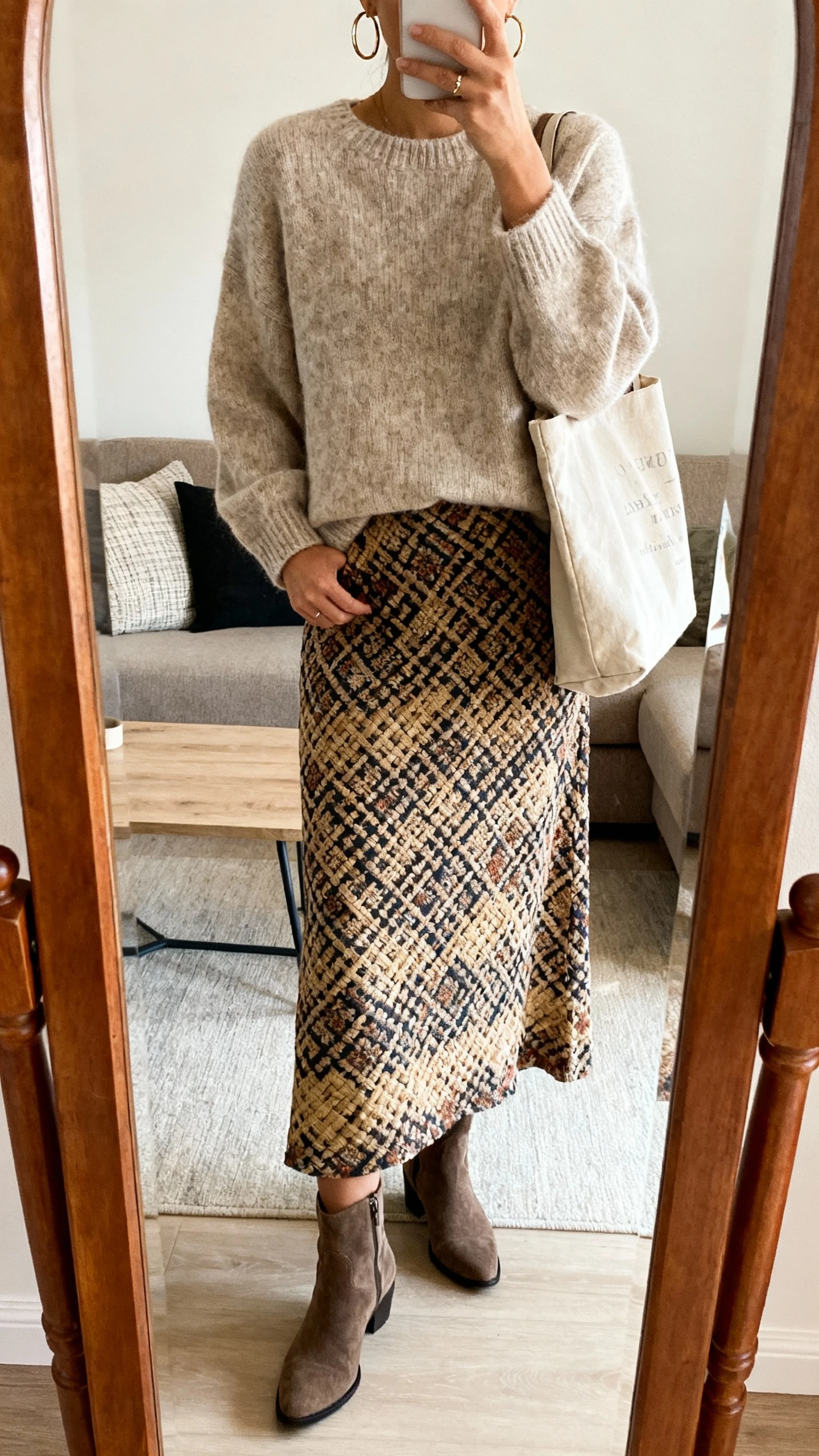 iPhone mirror selfie of a woman wearing a cozy pullover sweater with a printed woven maxi skirt, ankle boots, hoop earrings, and a casual tote; face not visible, cropped at neck, living room full-length mirror, warm indoor lighting, iPhone photo quality.