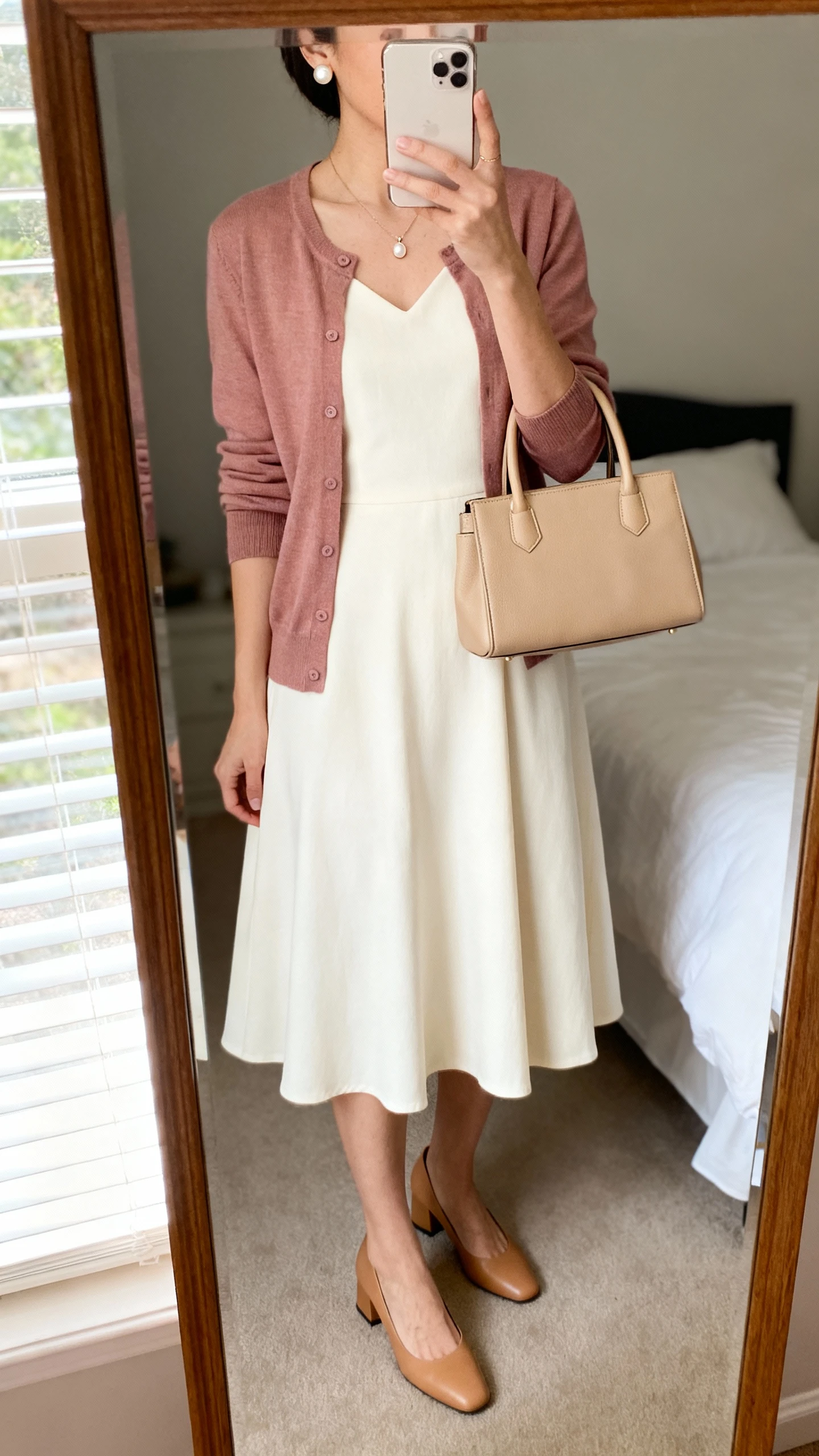 iPhone mirror selfie of a woman wearing a cream A-line midi dress with elbow sleeves, a lightweight cardigan in dusty rose, camel low block-heel pumps, pearl stud earrings, a dainty pendant necklace, and a structured beige top-handle bag, face not visible, cropped at neck, bedroom mirror, natural window light, iPhone photo quality.