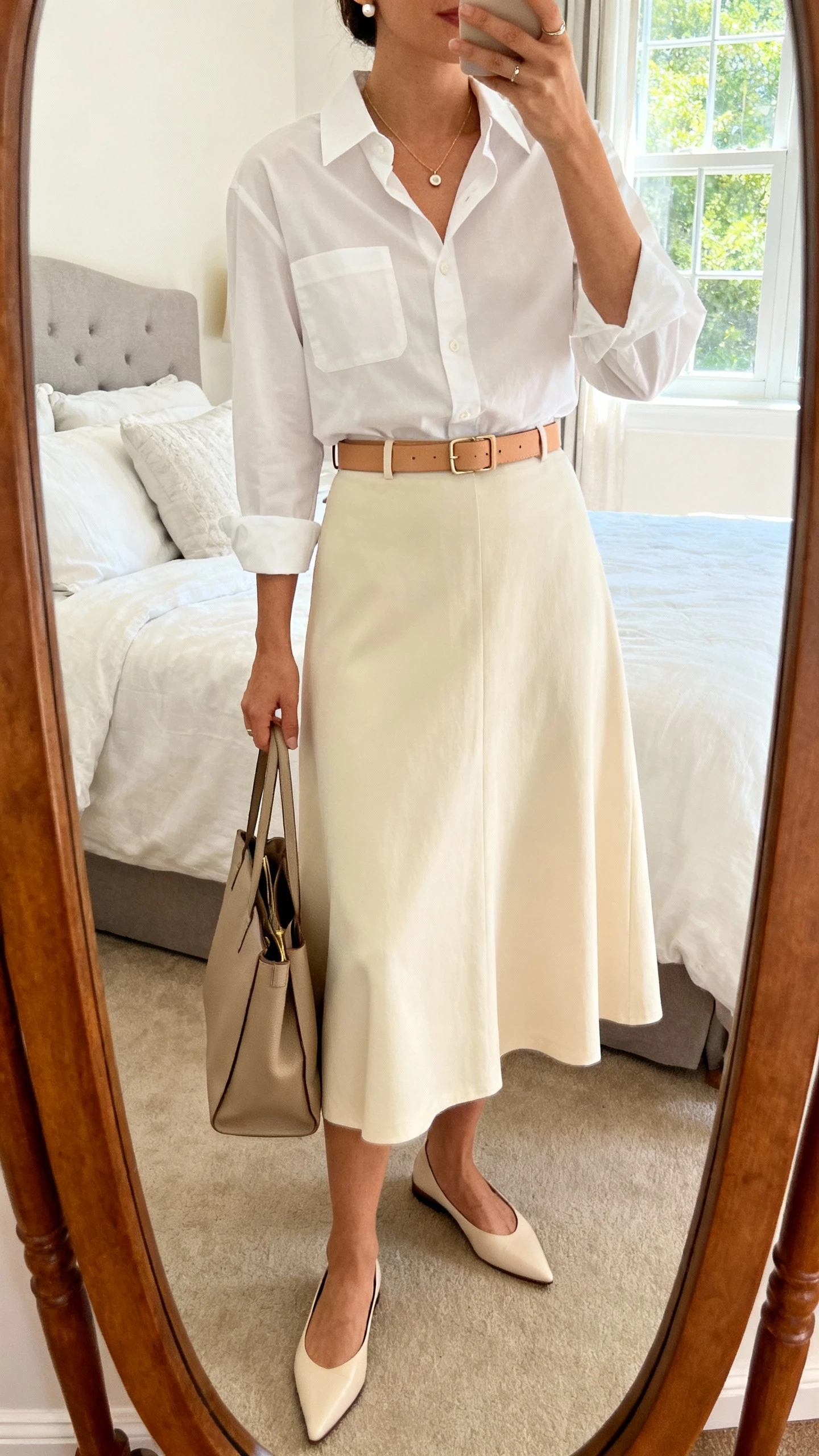iPhone mirror selfie of a woman wearing a cream A-line midi skirt, white crisp button-down shirt, slim tan belt, pointed-toe flats, delicate pendant necklace, small stud earrings, and a structured tote; face not visible, cropped at neck, bedroom mirror, natural window light, iPhone photo quality.