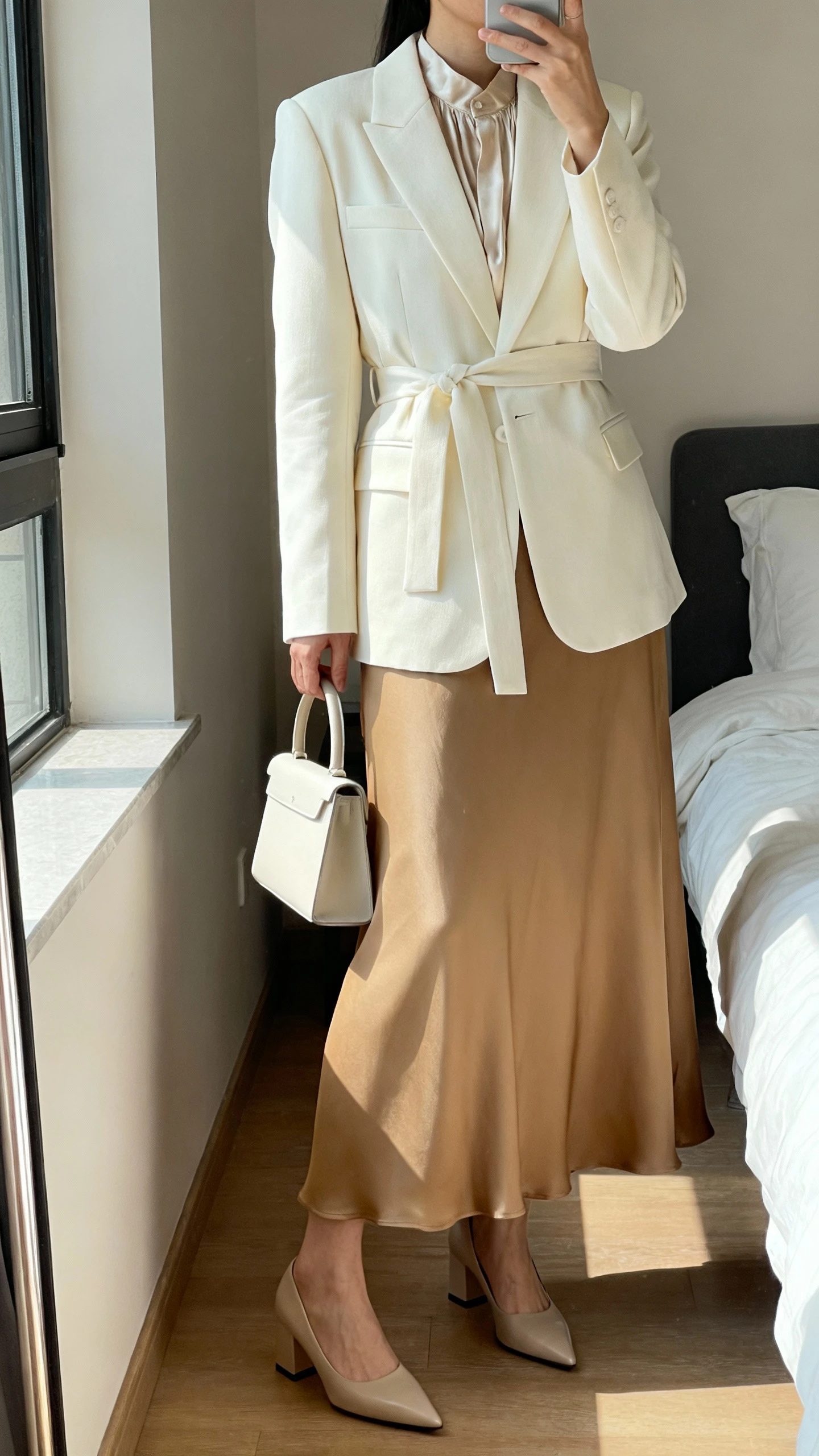 iPhone mirror selfie of a woman wearing a cream belted blazer over a modest silk blouse, fluid camel maxi skirt, pointed block-heel pumps, and a minimal top-handle bag, face not visible, cropped at neck, bedroom mirror, natural window light, iPhone photo quality.
