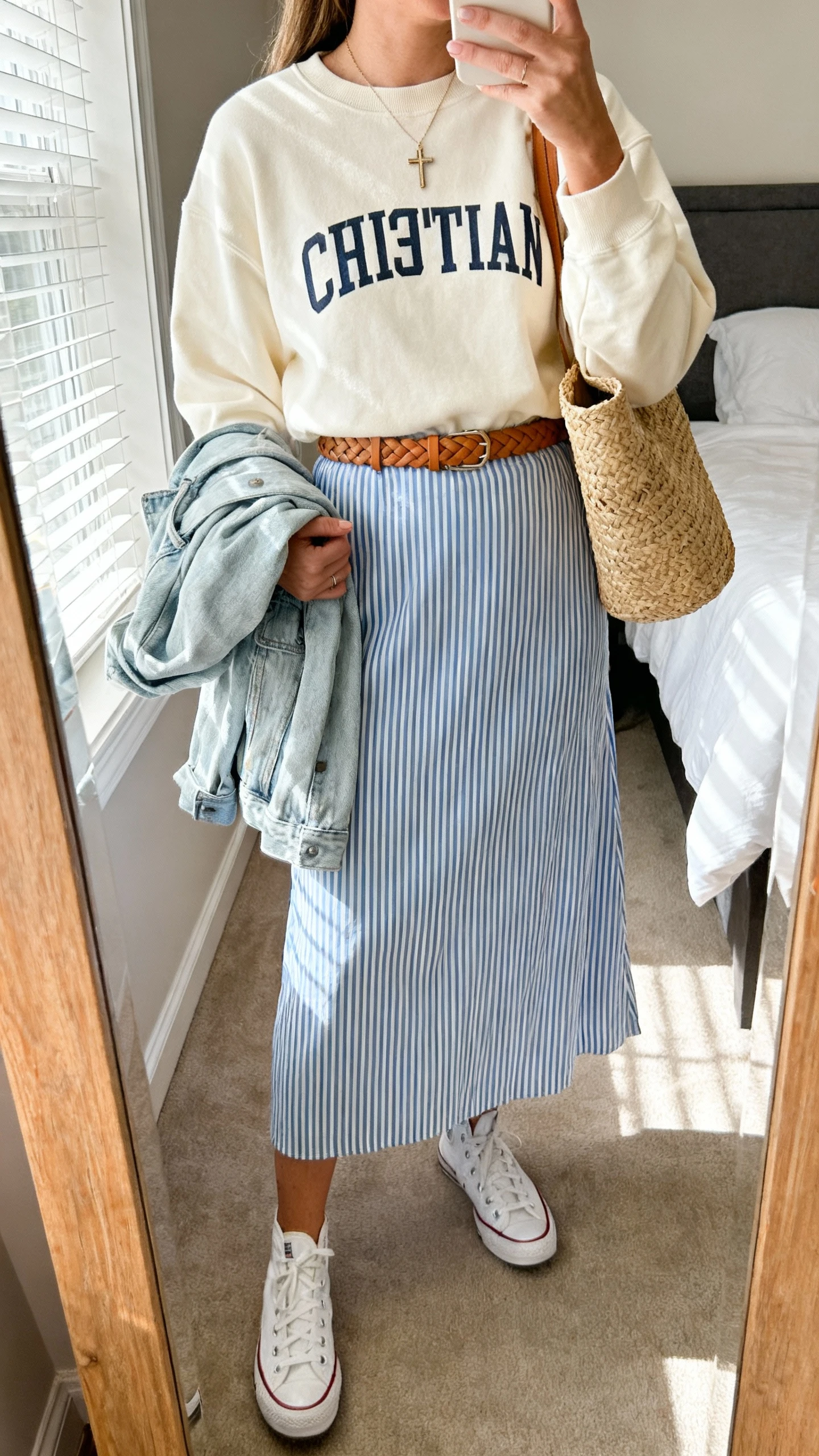 iPhone mirror selfie of a woman wearing a cream Christian crewneck layered over a blue-and-white striped midi skirt, tan braided belt, white canvas sneakers, straw tote, dainty cross pendant, and a light denim jacket draped over the arm, face not visible, bedroom mirror, soft afternoon window light, iPhone photo quality.