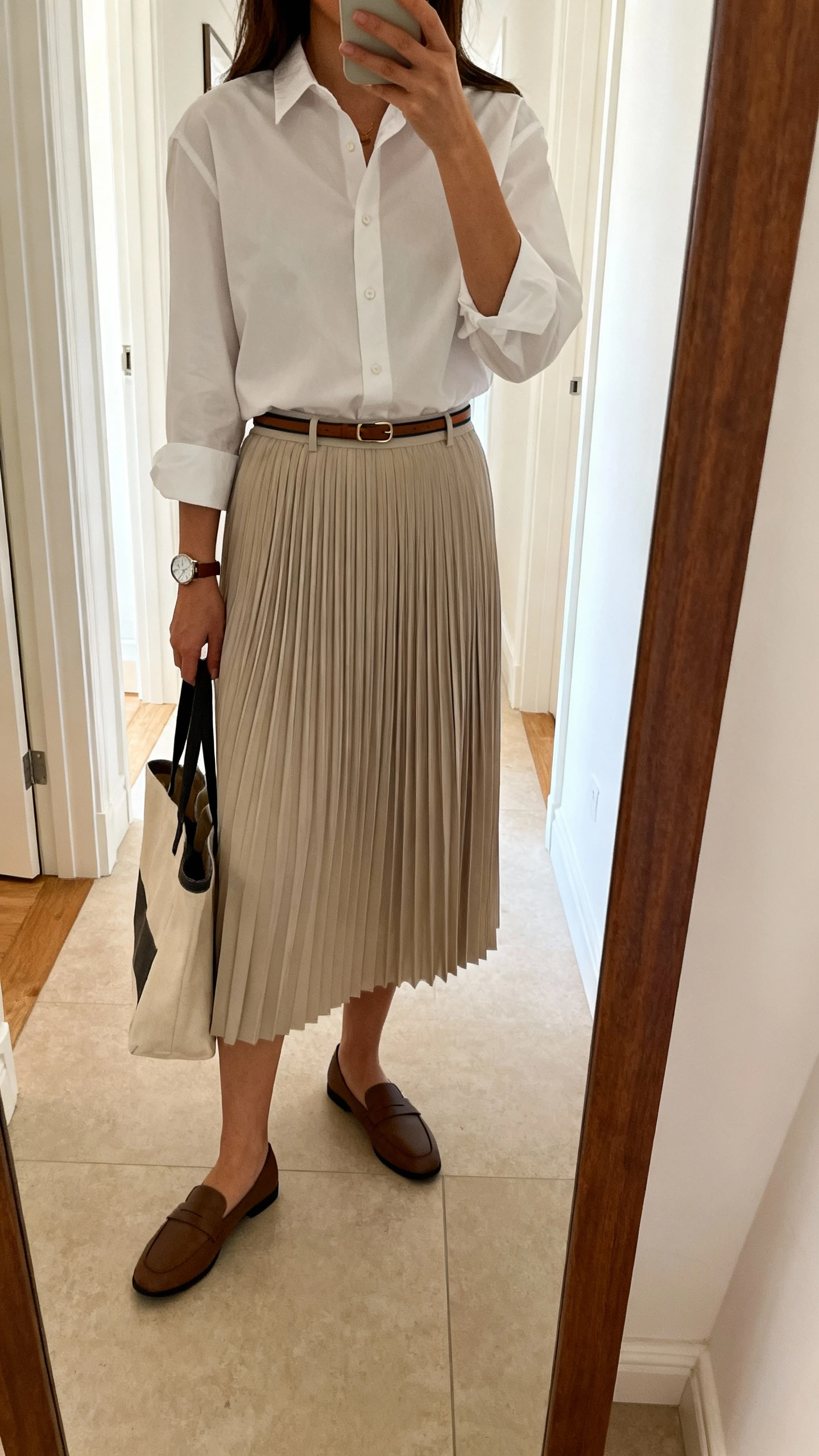 iPhone mirror selfie of a woman wearing a crisp button-down shirt tucked into a pleated midi skirt with a skinny belt, loafers, a classic watch, and a tote bag, face not visible, cropped at shoulders, full-length hallway mirror, natural daylight, iPhone photo quality.
