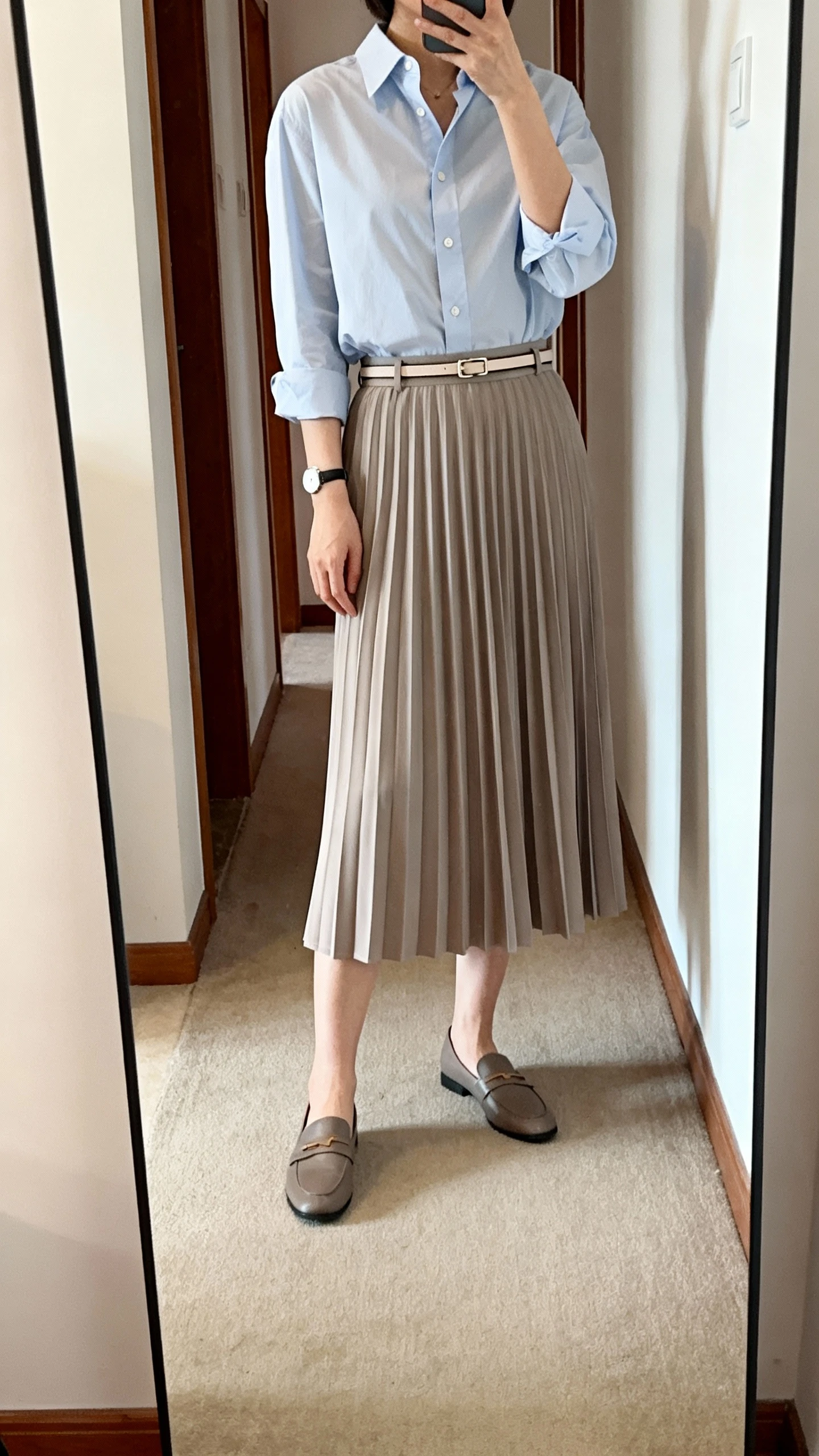 iPhone mirror selfie of a woman wearing a crisp button-up shirt tucked into a pleated midi skirt with a thin belt, loafers, and a simple watch, face not visible, cropped at shoulders, full-length mirror in hallway, natural daylight, iPhone photo quality.