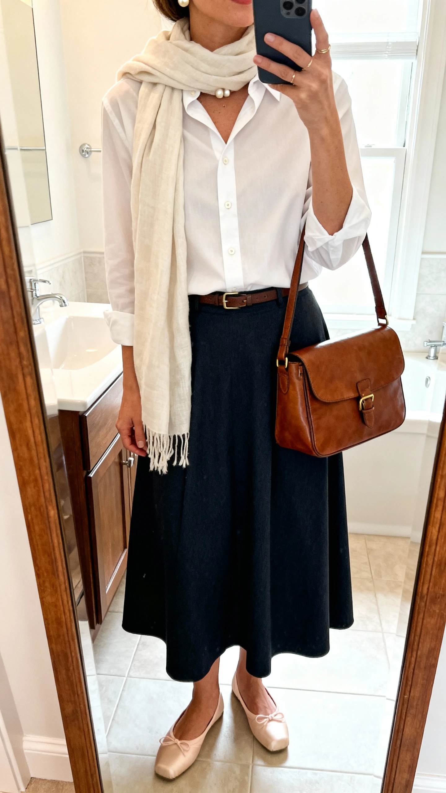 iPhone mirror selfie of a woman wearing a crisp button-up shirt tucked into a full midi skirt with a light scarf draped gently, classic ballet flats, pearl studs, and a leather satchel, face not visible, cropped at neck, bathroom mirror, bright indoor lighting, iPhone photo quality.