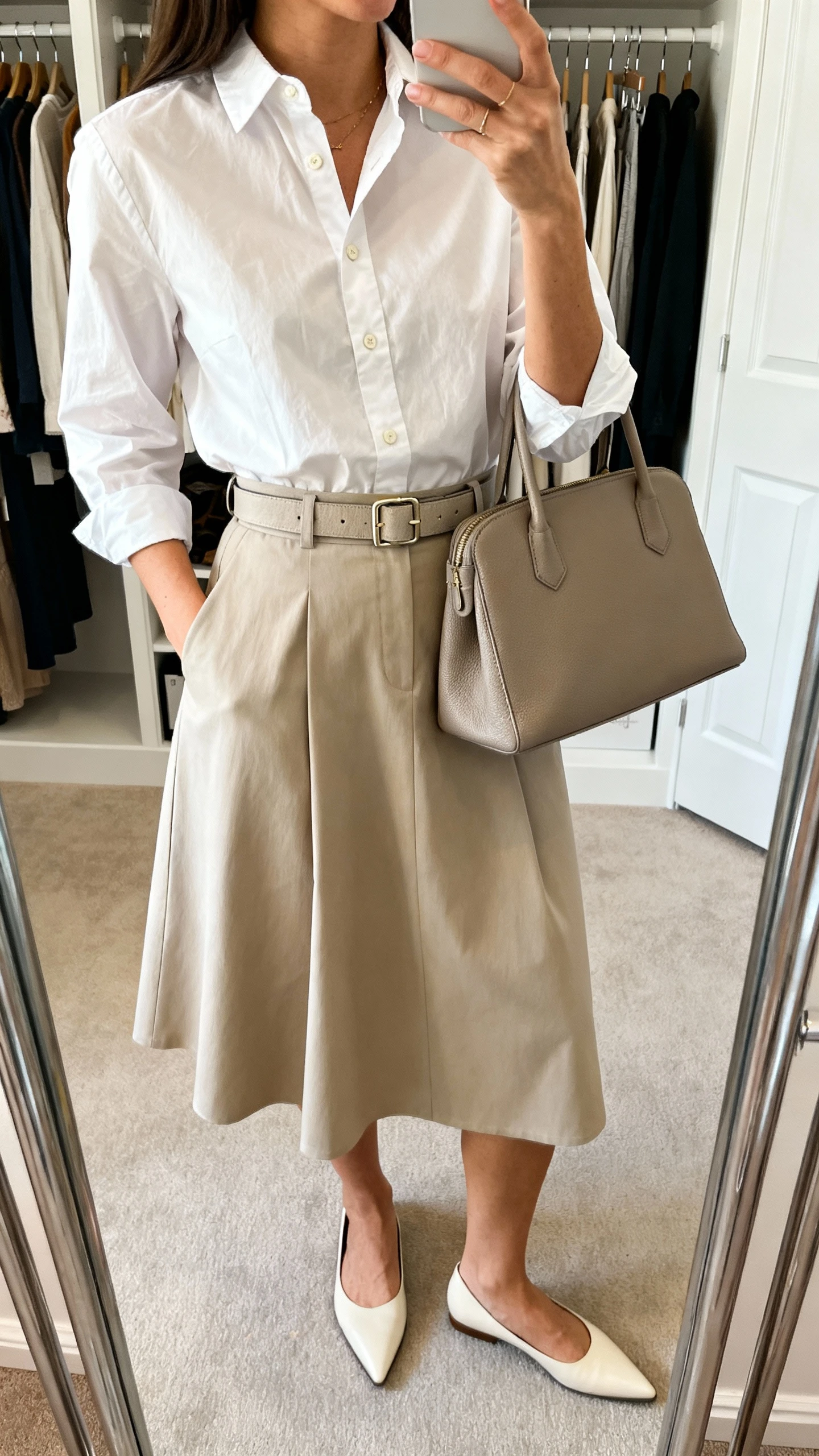 iPhone mirror selfie of a woman wearing a crisp buttoned-up poplin blouse tucked into a belted A-line skirt, structured tote and pointed flats, face not visible, cropped at neck, closet mirror, bright indoor lighting, iPhone photo quality.