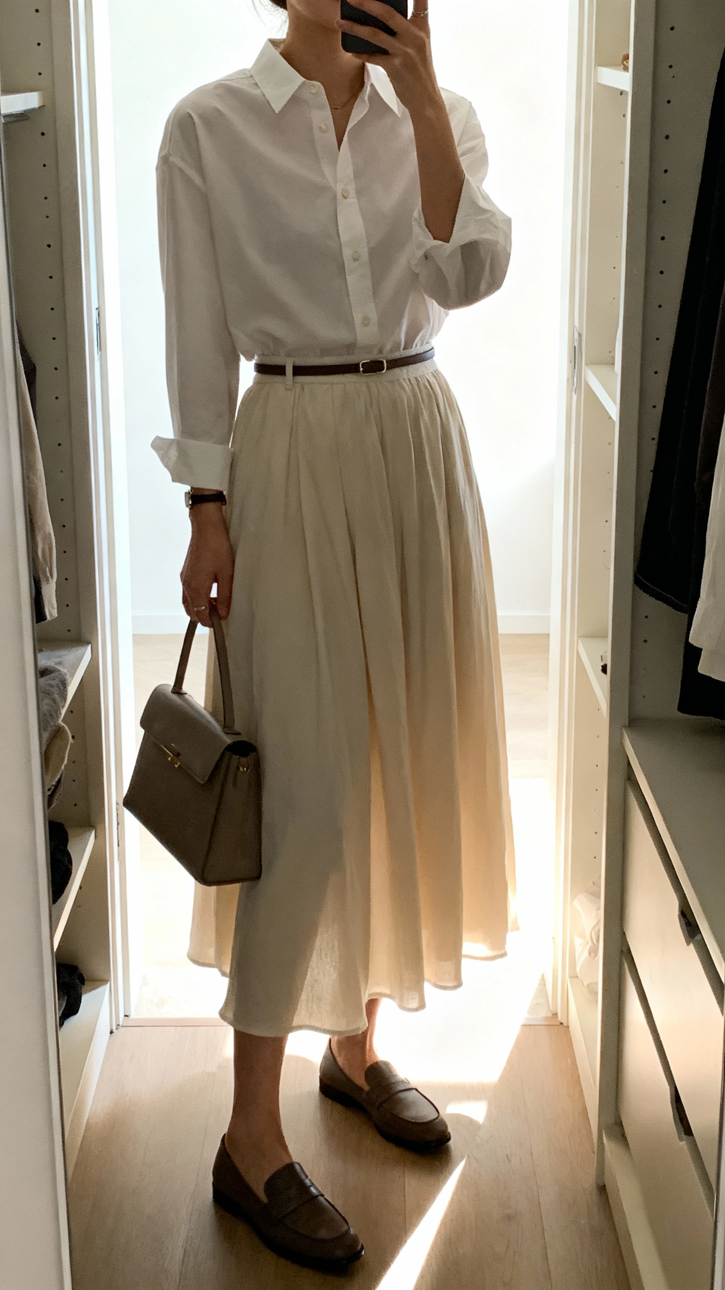 iPhone mirror selfie of a woman wearing a crisp button-up shirt tucked into a flowy maxi skirt, with a thin waist belt, loafers, and a structured tote, face not visible, cropped at neck, closet full-length mirror, bright indoor lighting, iPhone photo quality.