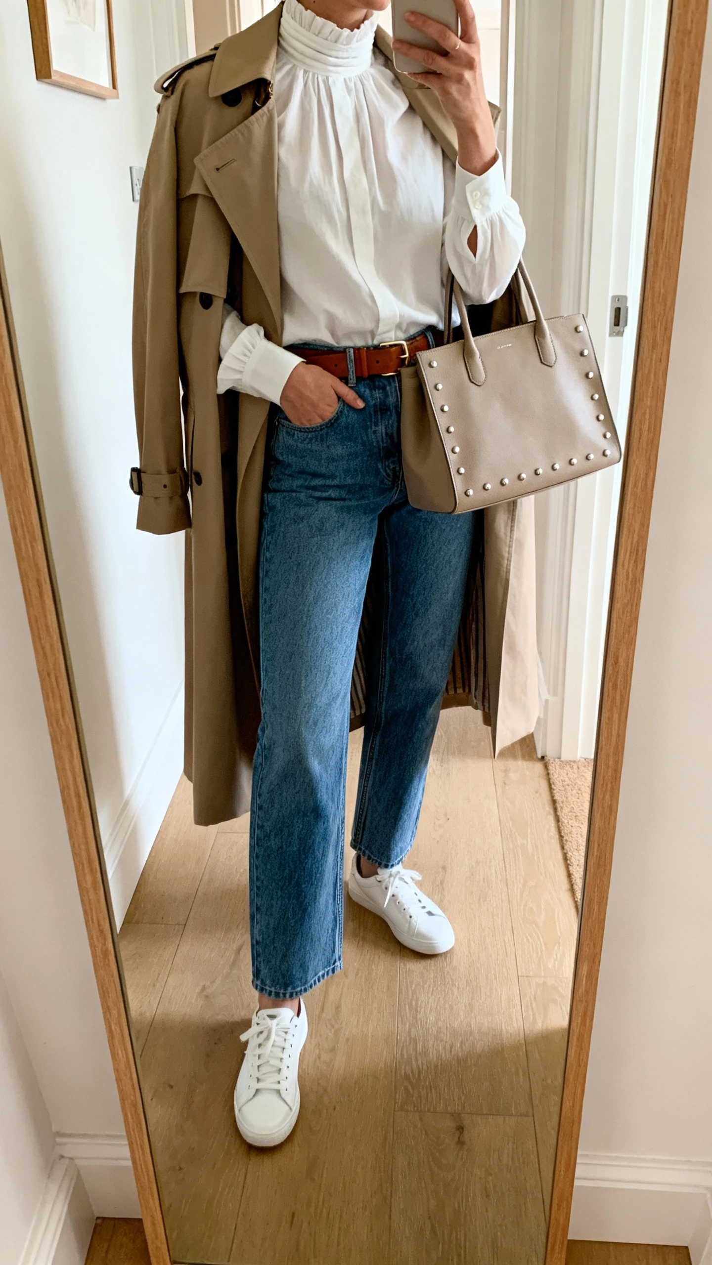 iPhone mirror selfie of a woman wearing a crisp high-neck blouse with straight-leg blue denim, a classic trench coat draped over shoulders, white sneakers, leather belt, and tote bag with simple studs; face not visible, cropped at neck, entryway mirror, natural daylight, iPhone photo quality.