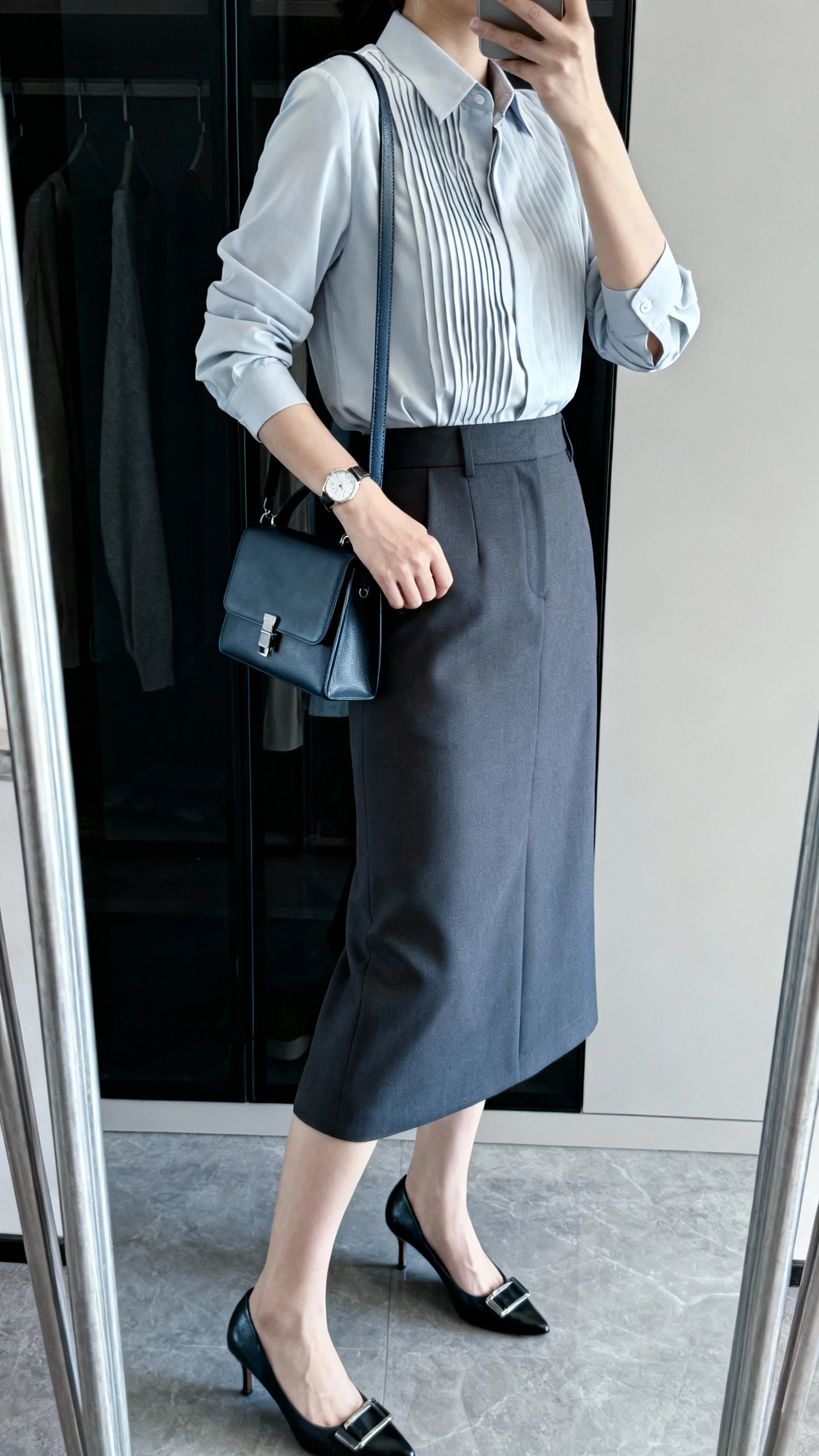iPhone mirror selfie of a woman wearing a crisp pintuck blouse tucked into a midi suiting skirt, with a minimalist watch, structured crossbody, and classic pumps, face not visible, side angle in closet mirror, cool indoor lighting, iPhone photo quality.