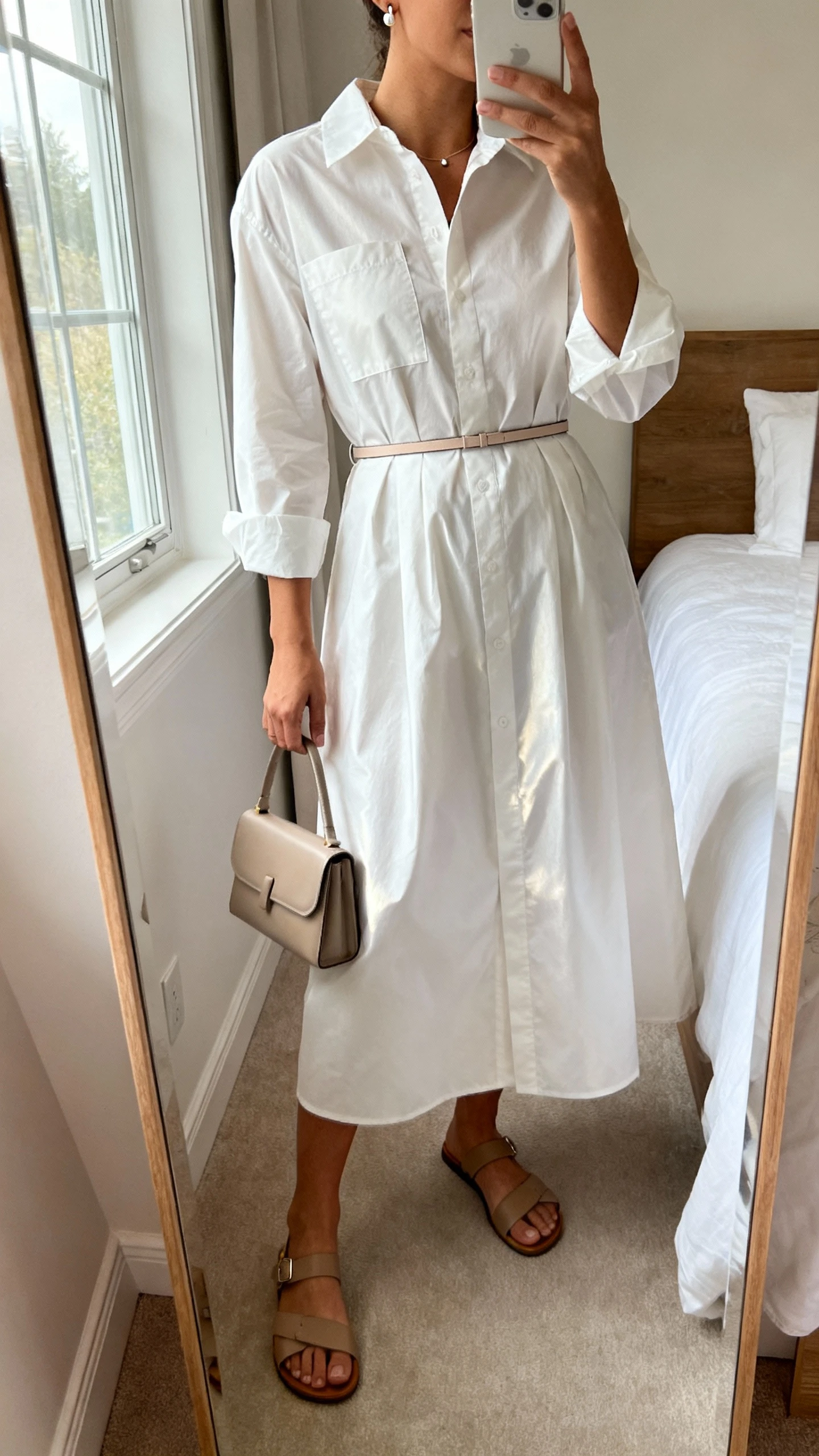 iPhone mirror selfie of a woman wearing a crisp poplin shirt dress with a subtle sheen, thin waist belt, delicate stud earrings, simple leather sandals, and a small structured tote, face not visible, cropped at neck, bedroom mirror, natural window light, iPhone photo quality.