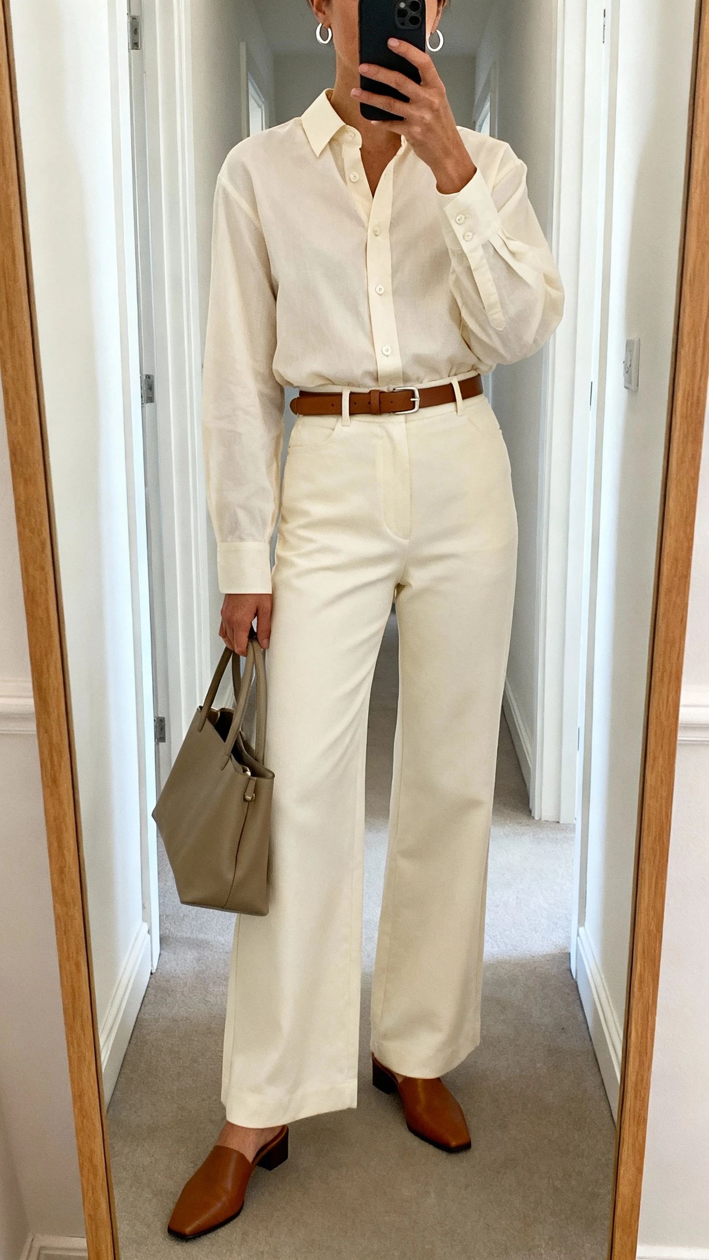 iPhone mirror selfie of a woman wearing a crisp poplin button-up with covered placket and high-rise wide-leg trousers (cream), polished mules, a slim leather belt, a structured tote, and small modern hoop earrings; face not visible, cropped at shoulders, full-length hallway mirror, natural daylight, iPhone photo quality.