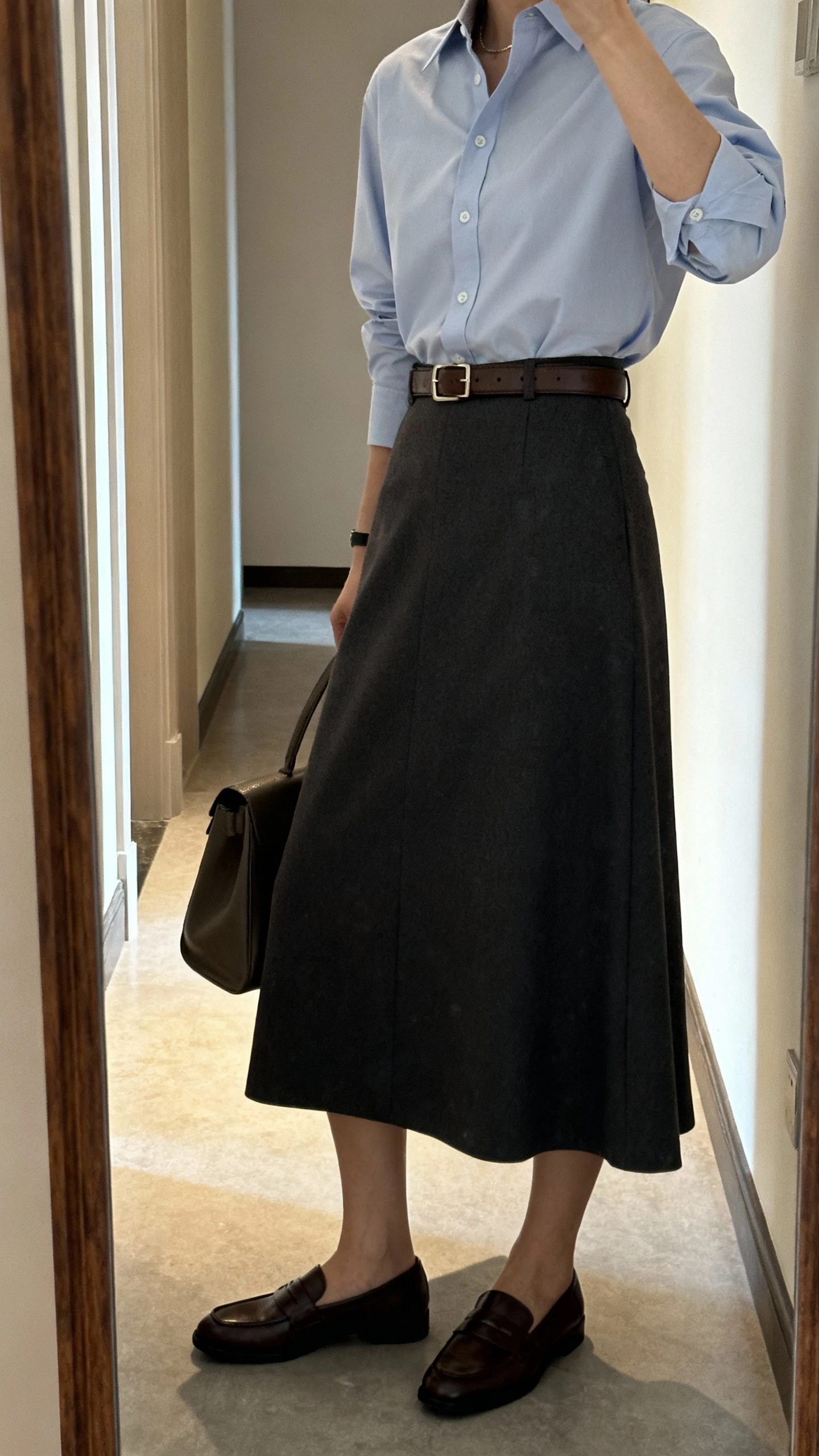 iPhone mirror selfie of a woman wearing a crisp tailored button-up shirt tucked into an A-line long skirt, slim leather belt, classic loafers, and a structured tote, face not visible, side angle in a hallway full-length mirror with natural indoor lighting, iPhone photo quality.