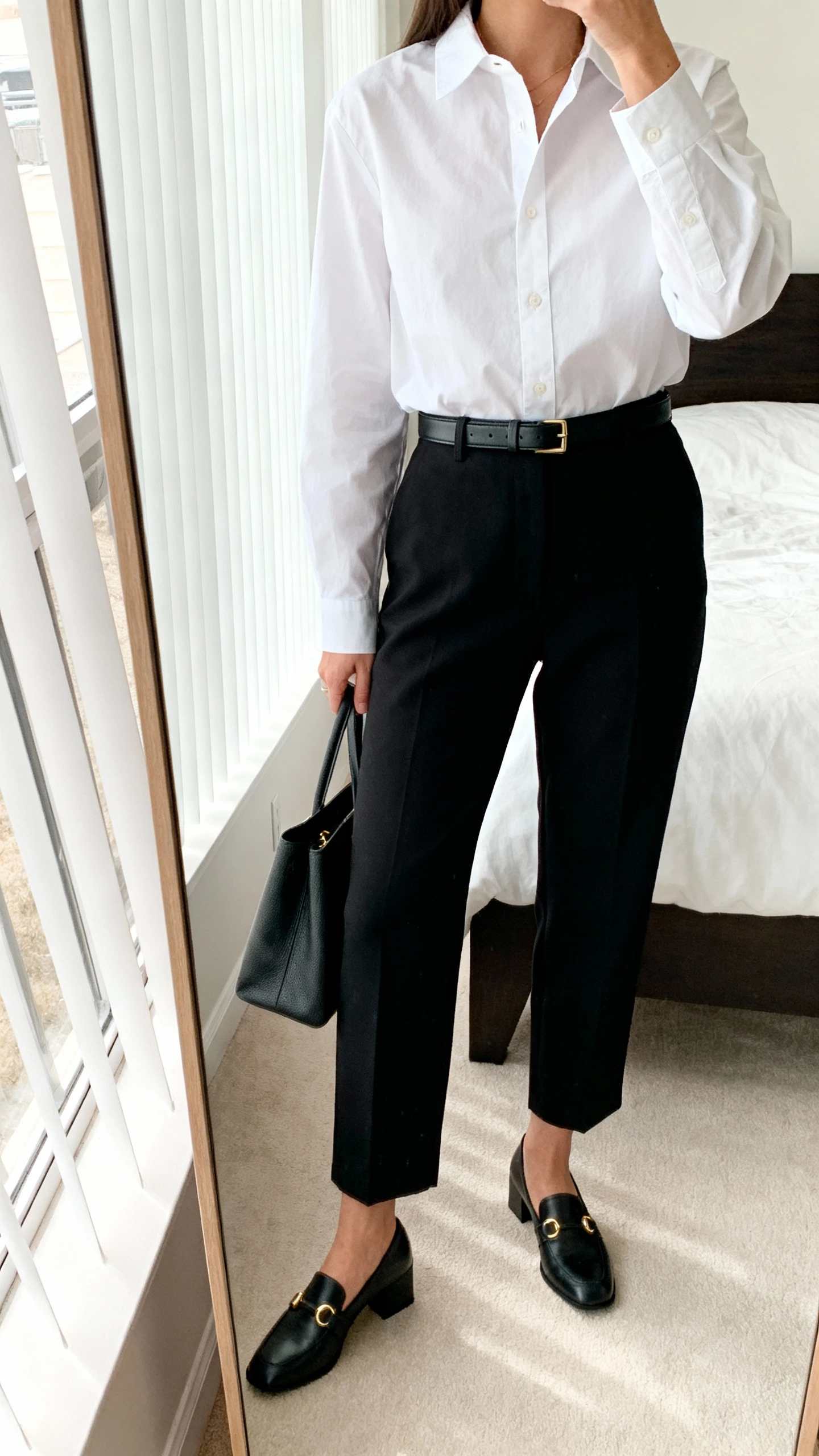 iPhone mirror selfie of a woman wearing a crisp white poplin button-up tucked into tailored black trousers with a slim leather belt, low block-heel loafers, minimalist gold studs, and a structured tote, face not visible, cropped at neck, bedroom mirror, natural window light, iPhone photo quality.