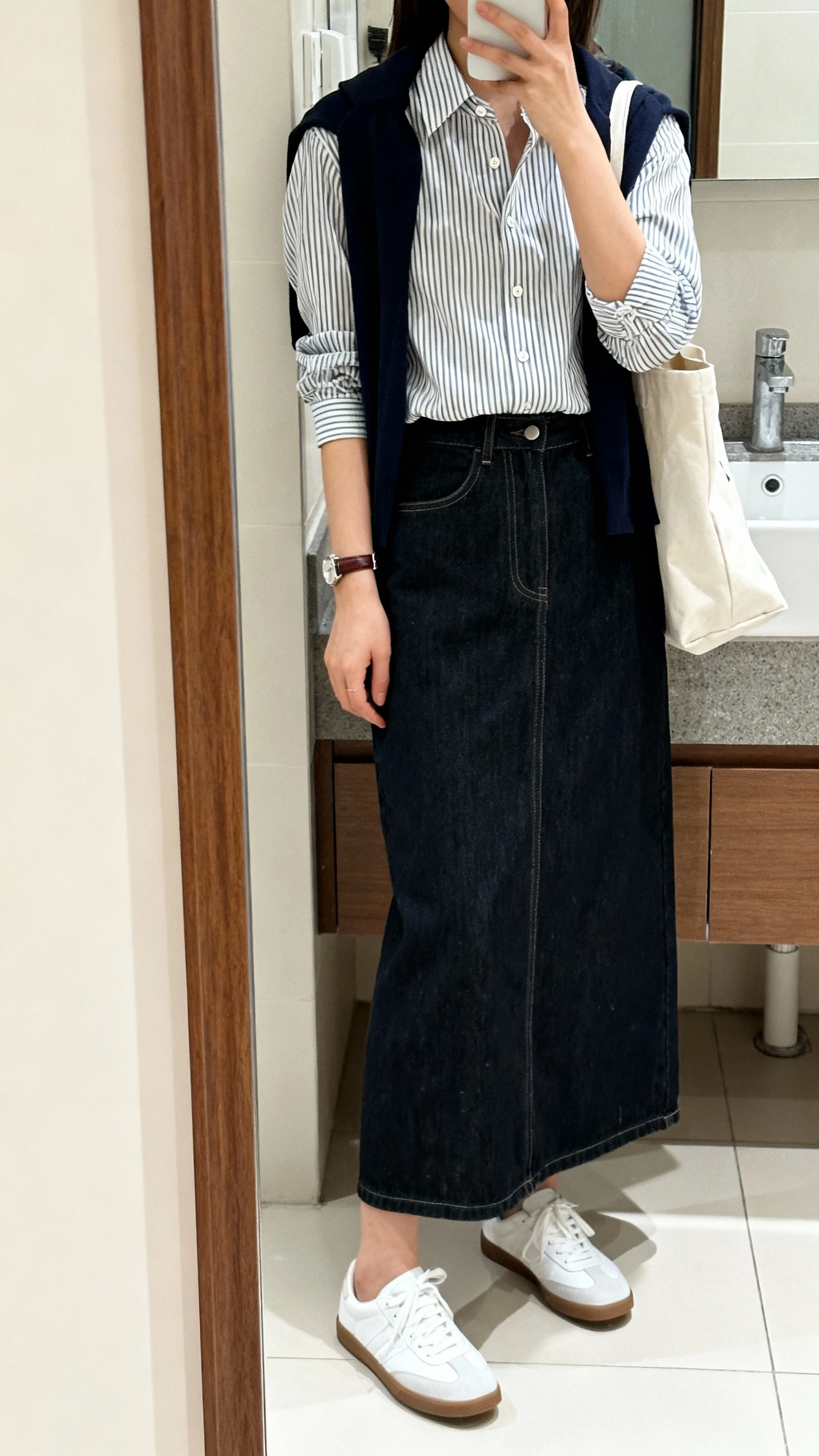 iPhone mirror selfie of a woman wearing a dark denim maxi skirt with a tucked-in striped button-up, slim cardigan tied over shoulders, white sneakers, leather watch, and a tote bag, face not visible, cropped at shoulders, bathroom full-length mirror, bright indoor lighting, iPhone photo quality.