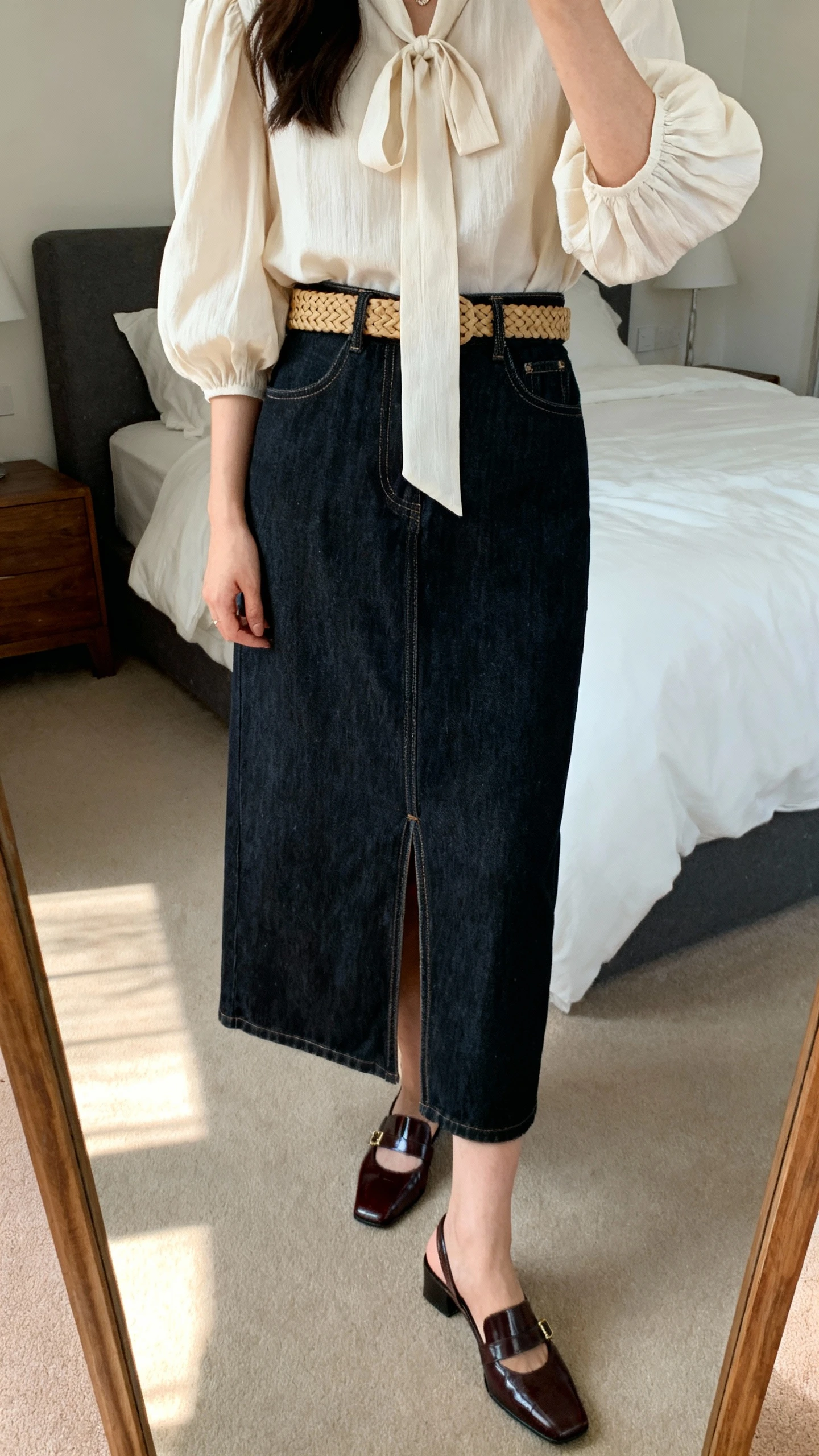 iPhone mirror selfie of a woman wearing a dark-wash denim maxi skirt with a modest front slit, a feminine blouse with puff sleeves and a tie-neck, polished closed-toe mules, and a woven belt, face not visible, cropped at shoulders, bedroom mirror, afternoon natural light, iPhone photo quality.