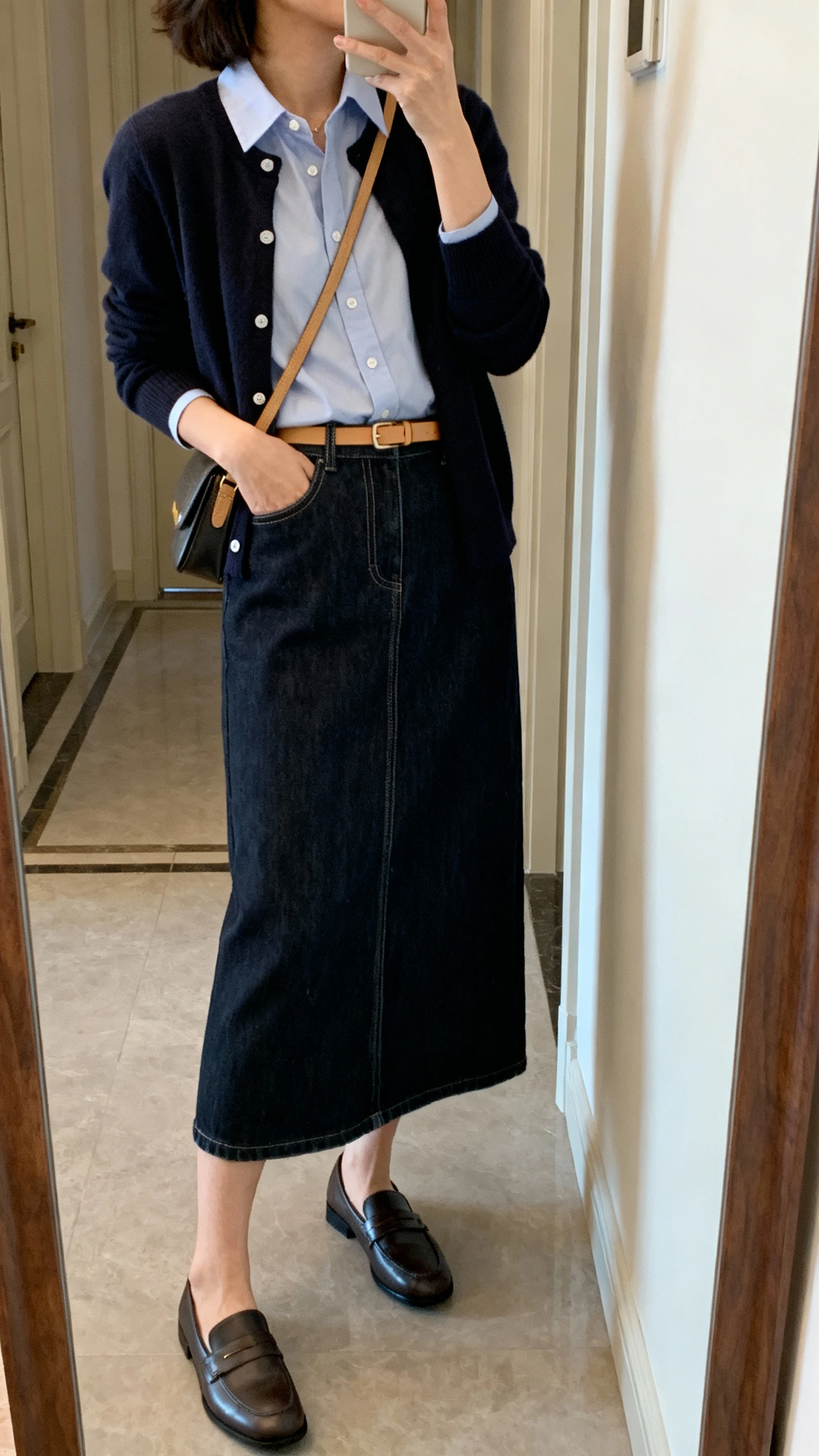 iPhone mirror selfie of a woman wearing a dark-wash denim midi skirt with a crisp button-up shirt, a cardigan layered on top, comfortable loafers, a crossbody bag, and a thin tan belt; face not visible, side angle, entryway full-length mirror, soft indoor lighting, iPhone photo quality.