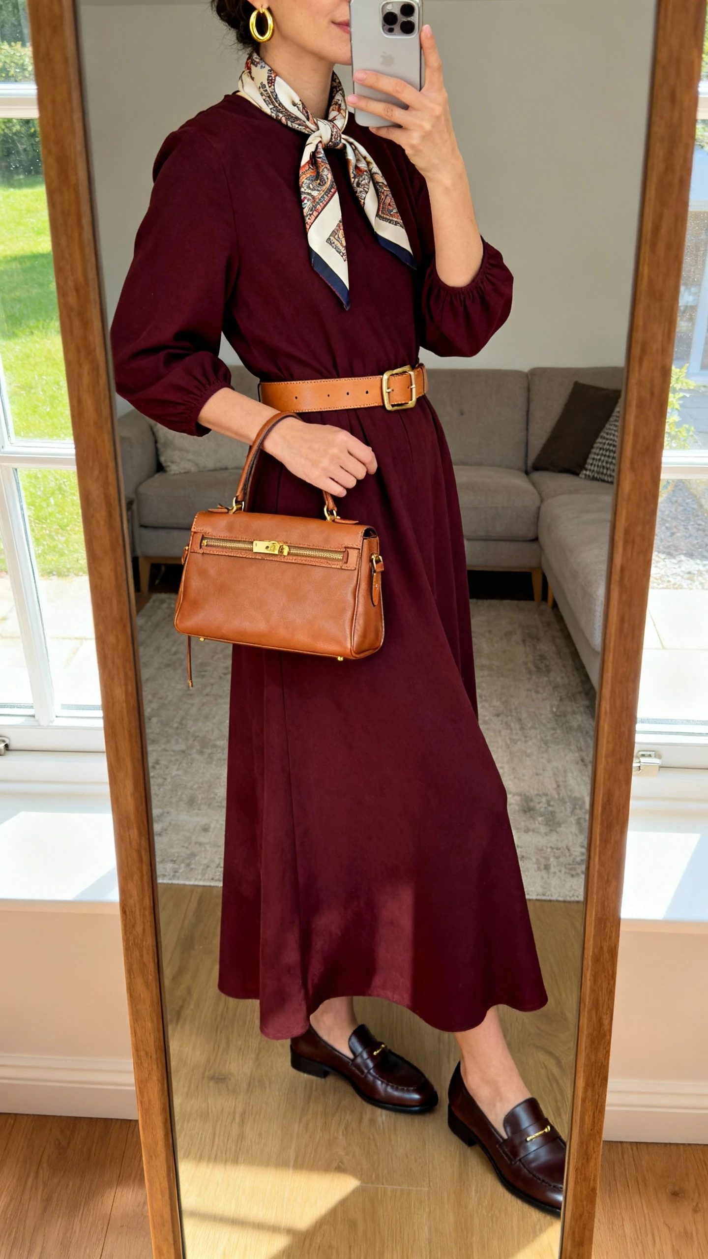 iPhone mirror selfie of a woman wearing a deep burgundy maxi dress styled with heritage accessories: a camel leather belt, vintage-inspired silk scarf tied at the neck, dark brown loafers, a structured tan satchel, and gold hoop earrings, face not visible, side angle, living room mirror, afternoon window light, iPhone photo quality.