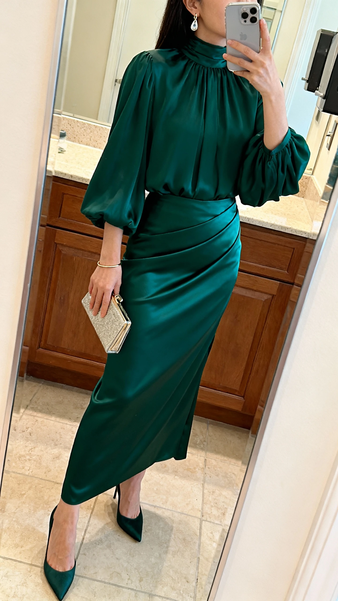 iPhone mirror selfie of a woman wearing a deep emerald bias-cut satin maxi skirt, flowy long-sleeve high-neck blouse, sleek kitten heels, compact clutch, elegant drop earrings, and a thin bracelet, face not visible, side angle, bathroom mirror, warm indoor lighting, iPhone photo quality.