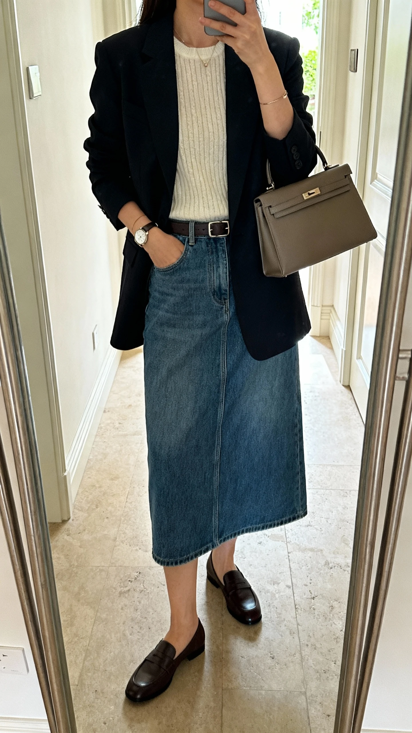iPhone mirror selfie of a woman wearing a denim midi skirt with a refined knit top, tailored blazer, loafers, slim belt, delicate watch, and a medium satchel, face not visible, cropped at neck, entryway full-length mirror, natural morning light, iPhone photo quality.