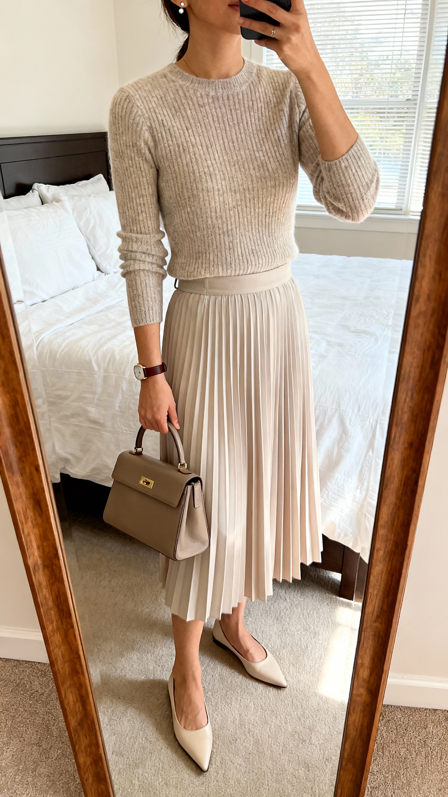 iPhone mirror selfie of a woman wearing a fine-knit fitted sweater tucked into a softly pleated midi skirt with pointed-toe flats, minimal stud earrings, a slim leather watch, and a structured top-handle bag, face not visible, cropped at neck, bedroom mirror, natural window light, iPhone photo quality.