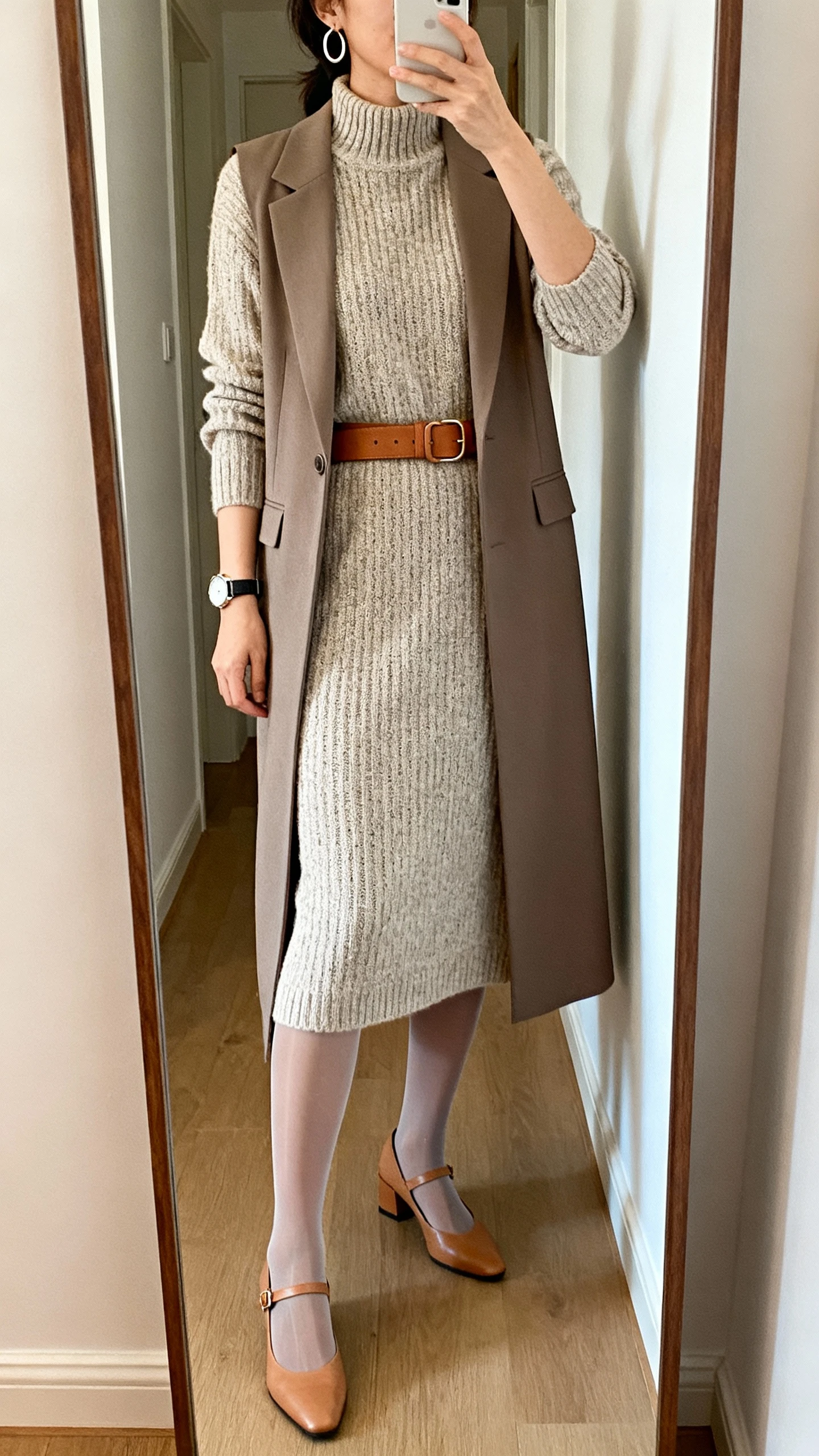 iPhone mirror selfie of a woman wearing a fine-knit midi dress with a mock neck, leather belt in tan, slingback flats, tailored longline vest, minimal hoop earrings, watch, and sheer tights, face not visible, cropped at shoulders, full-length mirror in hallway, soft indoor lighting, iPhone photo quality.