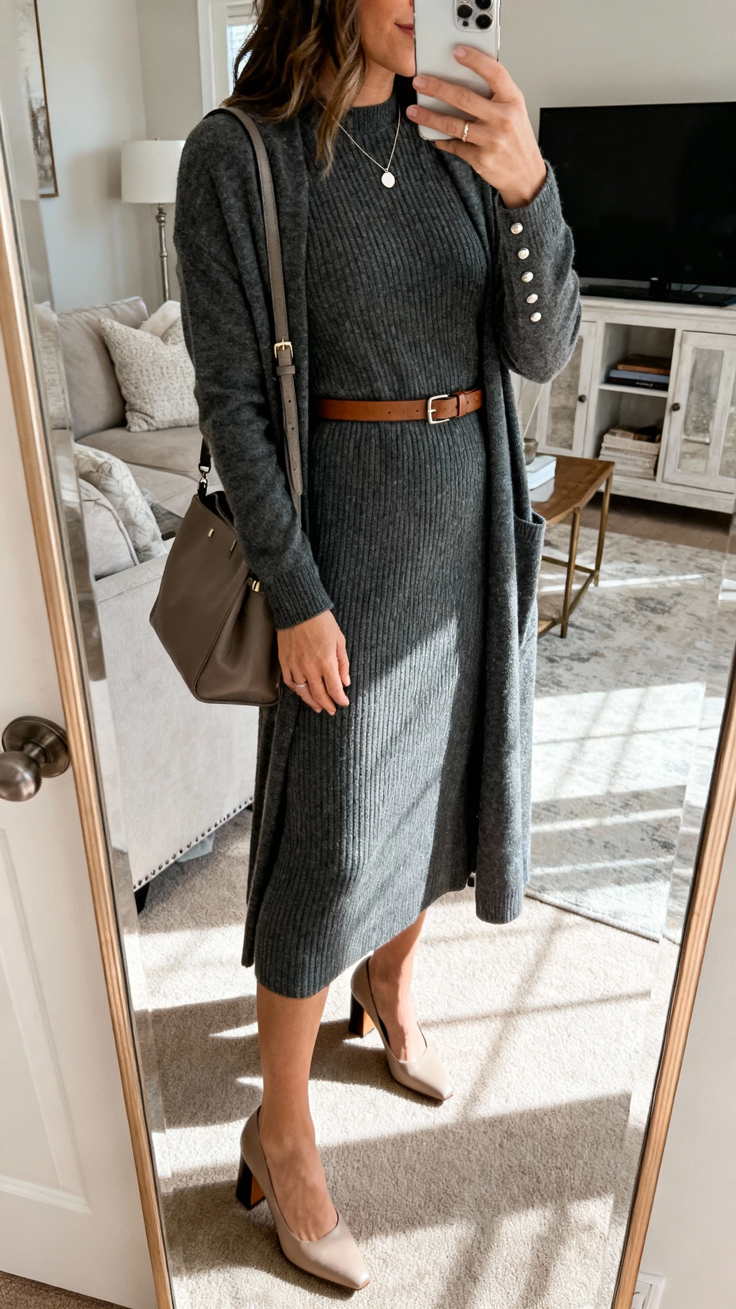 iPhone mirror selfie of a woman wearing a fine-knit midi dress in charcoal with a slim leather belt, block-heel pumps, a longline cardigan, studs with a minimal pendant, and a medium satchel, face not visible, side angle, living room full-length mirror with indoor morning light, iPhone photo quality.