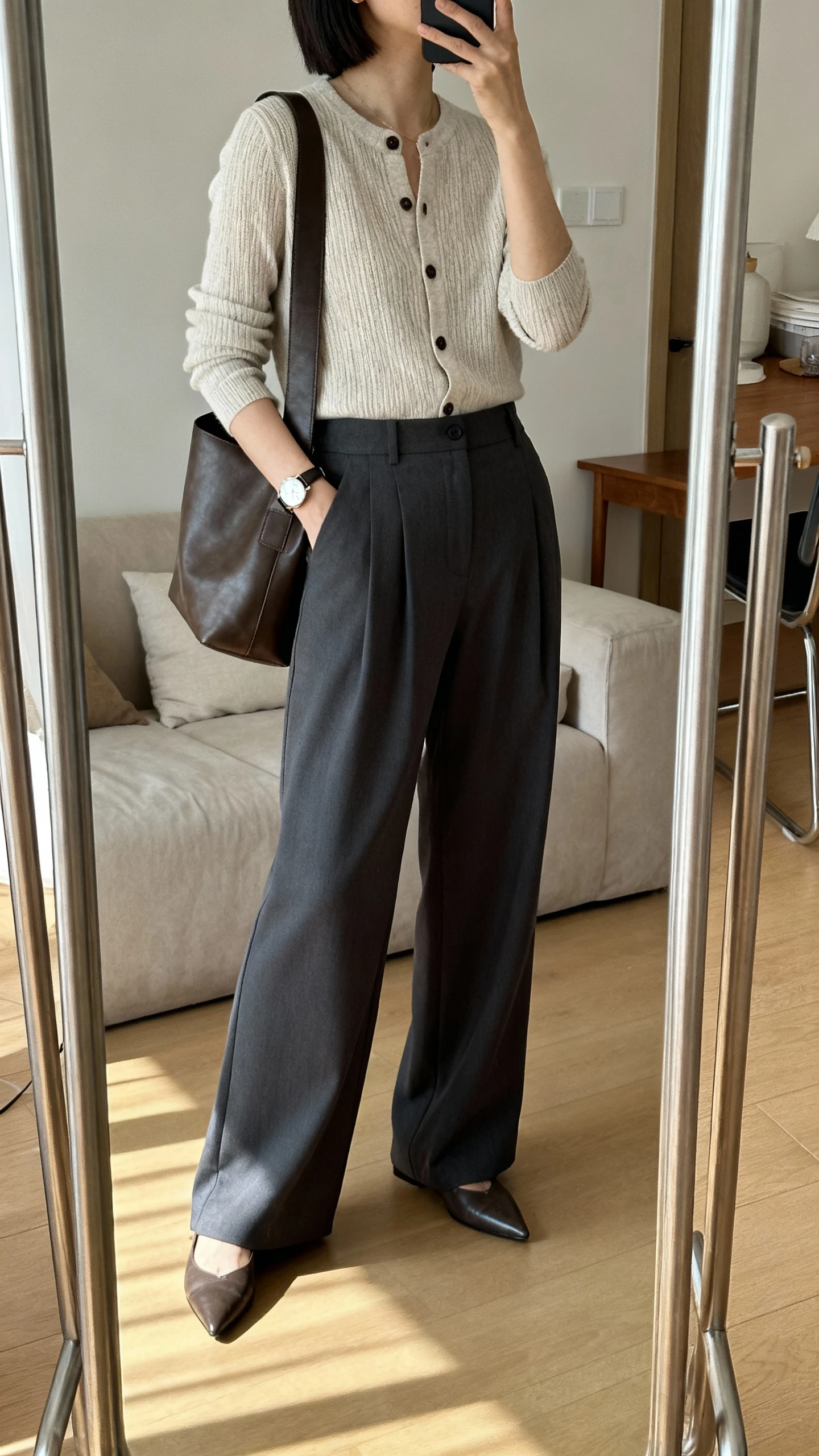 iPhone mirror selfie of a woman wearing a fine-knit cardigan buttoned as a top with high-waisted wide-leg trousers in charcoal, pointed-toe flats, a watch, and a leather shoulder bag, face not visible, side angle, living room full-length mirror, gentle afternoon indoor light, iPhone photo quality.