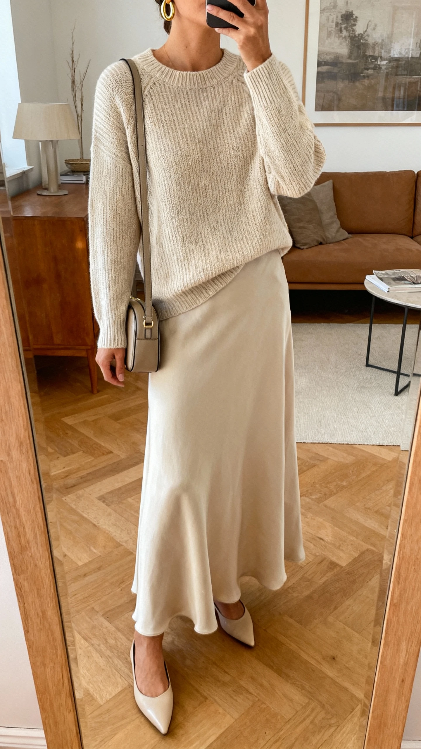 iPhone mirror selfie of a woman wearing a fine-knit crewneck sweater with a matching flowy maxi skirt, pointed flats, simple gold hoop earrings, and a compact shoulder bag, face not visible, cropped at neck, living-room mirror, warm indoor lighting, iPhone photo quality.