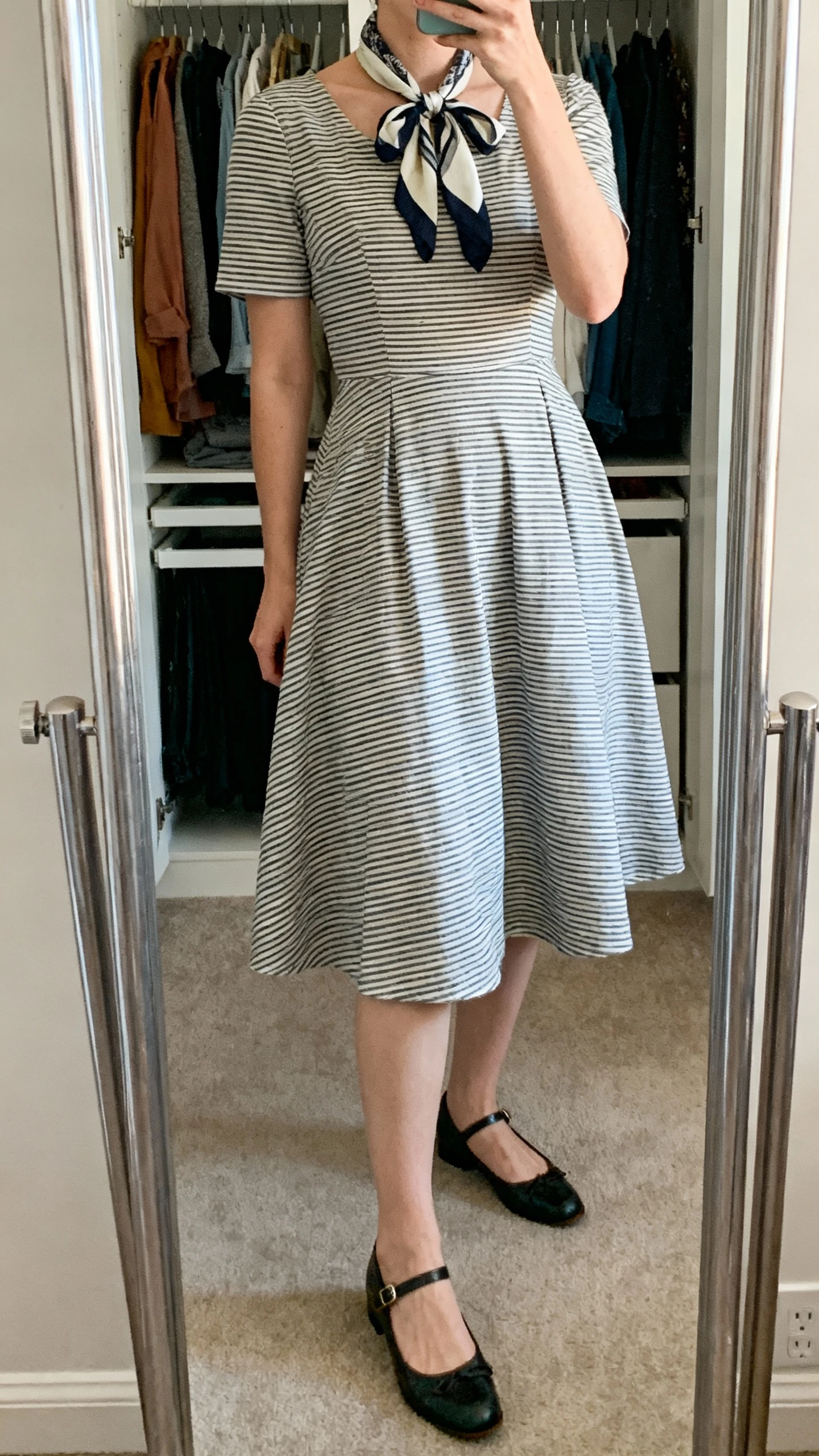 iPhone mirror selfie of a woman wearing a fit-and-flare striped dress with Mary Janes and a bow-tied scarf at the neck, face not visible, cropped at neck, closet mirror with gentle indoor lighting, iPhone photo quality.