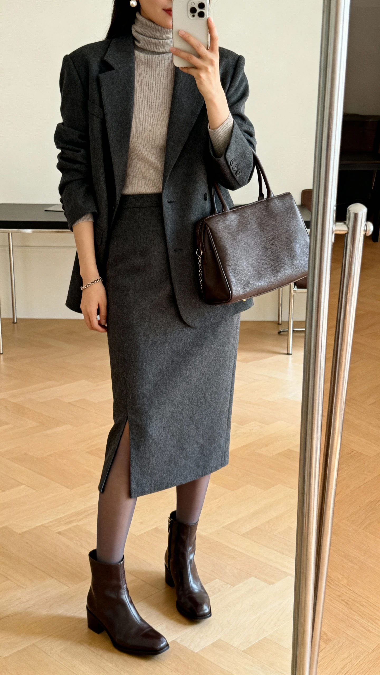 iPhone mirror selfie of a woman wearing a fitted cashmere-blend turtleneck, a wool midi pencil skirt with a back vent, a tailored charcoal blazer, polished ankle boots with opaque tights, a structured leather tote, and subtle pearl studs with a thin chain bracelet; face not visible, cropped at shoulders, full-length mirror, warm indoor lighting, iPhone photo quality.