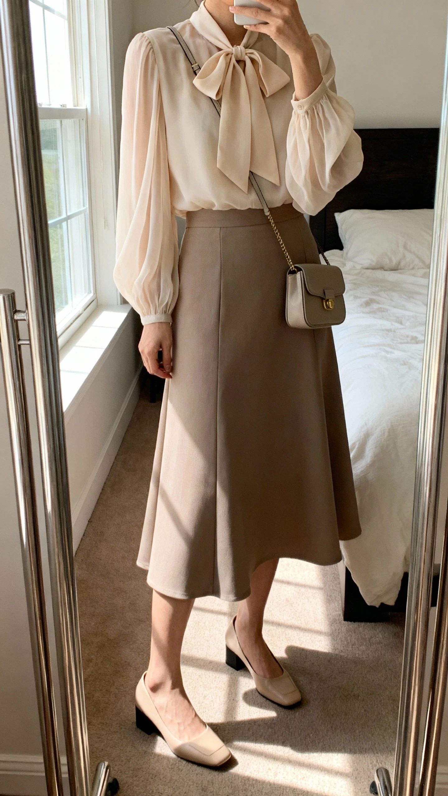 iPhone mirror selfie of a woman wearing a floaty bow-tie blouse with long sleeves and a soft drape, paired with a midi A-line skirt, low block-heel pumps, and a small crossbody bag; face not visible, cropped at neck, bedroom mirror, natural window light, iPhone photo quality.