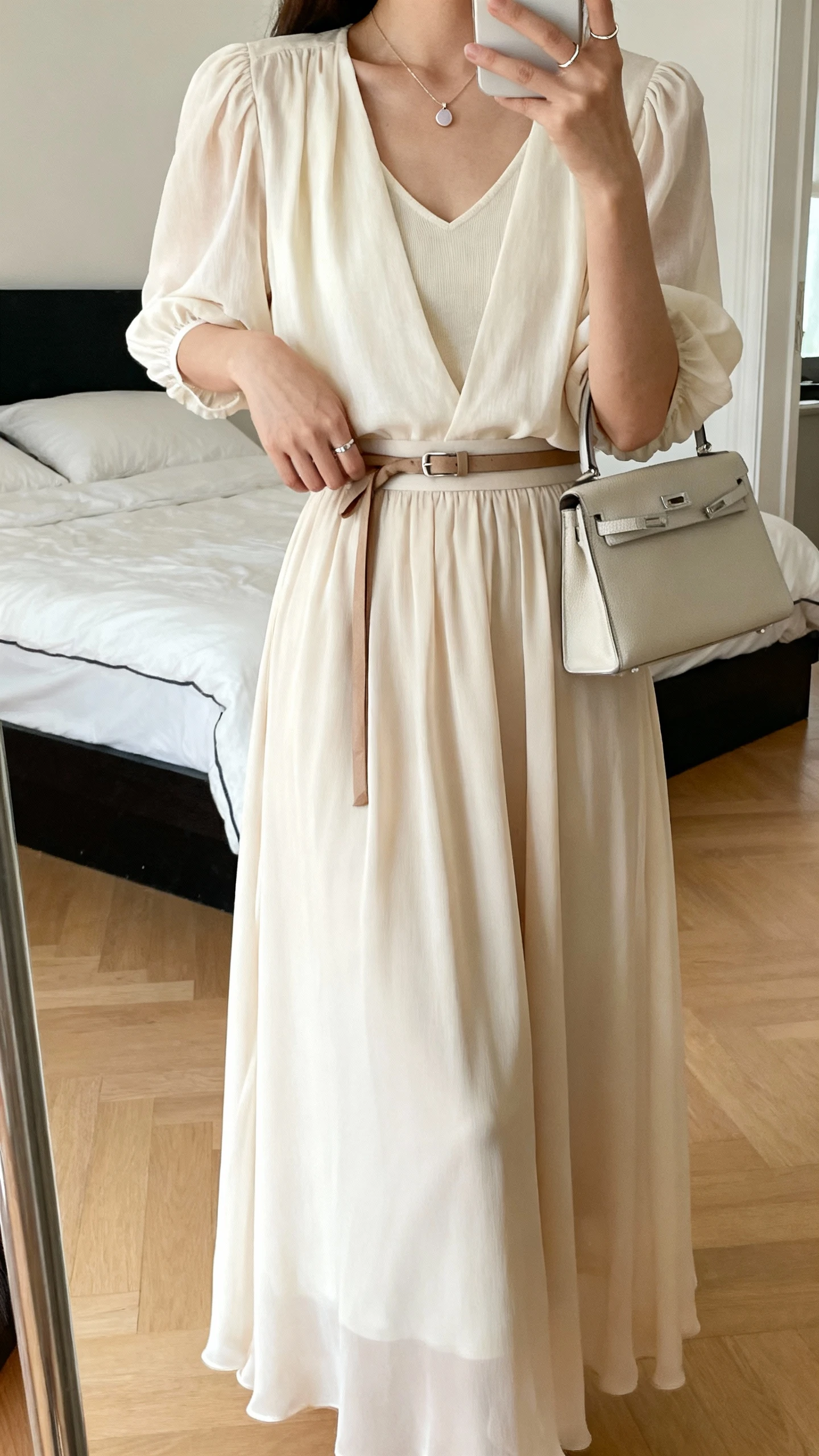 iPhone mirror selfie of a woman wearing a floaty maxi skirt in chiffon/georgette/crepe (solid or subtle print), a soft blouse with gathered sleeves or a gentle V over a camisole, pointed-toe flats or low-heeled slingbacks, a slim belt or self-tie at the waist, a light pendant necklace and a dainty ring, carrying a small handbag; face not visible, cropped at shoulders, bedroom mirror, natural daylight, iPhone photo quality.