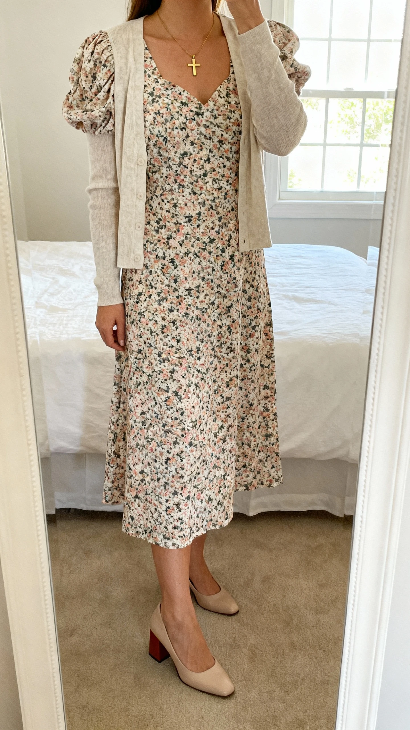 iPhone mirror selfie of a woman wearing a floral midi dress with a modest neckline and puff sleeves, a lightweight cardigan, closed-toe block-heel pumps, and a delicate gold pendant or small cross necklace, face not visible, cropped at neck, bedroom mirror, natural window light, iPhone photo quality.
