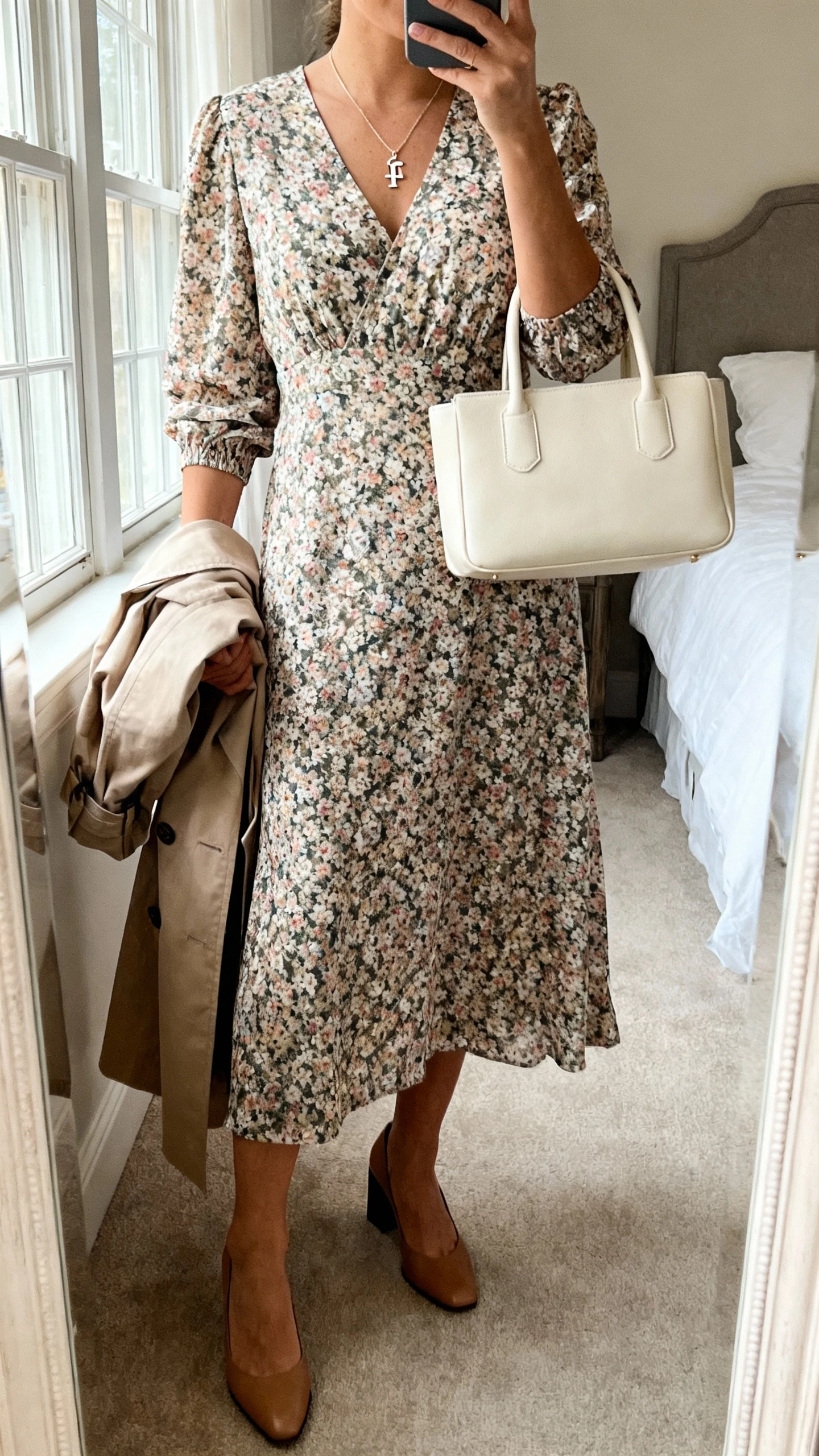 iPhone mirror selfie of a woman wearing a floral midi dress with elbow-length sleeves, low block-heel pumps, a delicate pendant necklace with a subtle faith symbol, a structured top-handle bag in cream, and a lightweight trench draped over her arm; face not visible, cropped at neck, bedroom mirror, natural window light, iPhone photo quality.