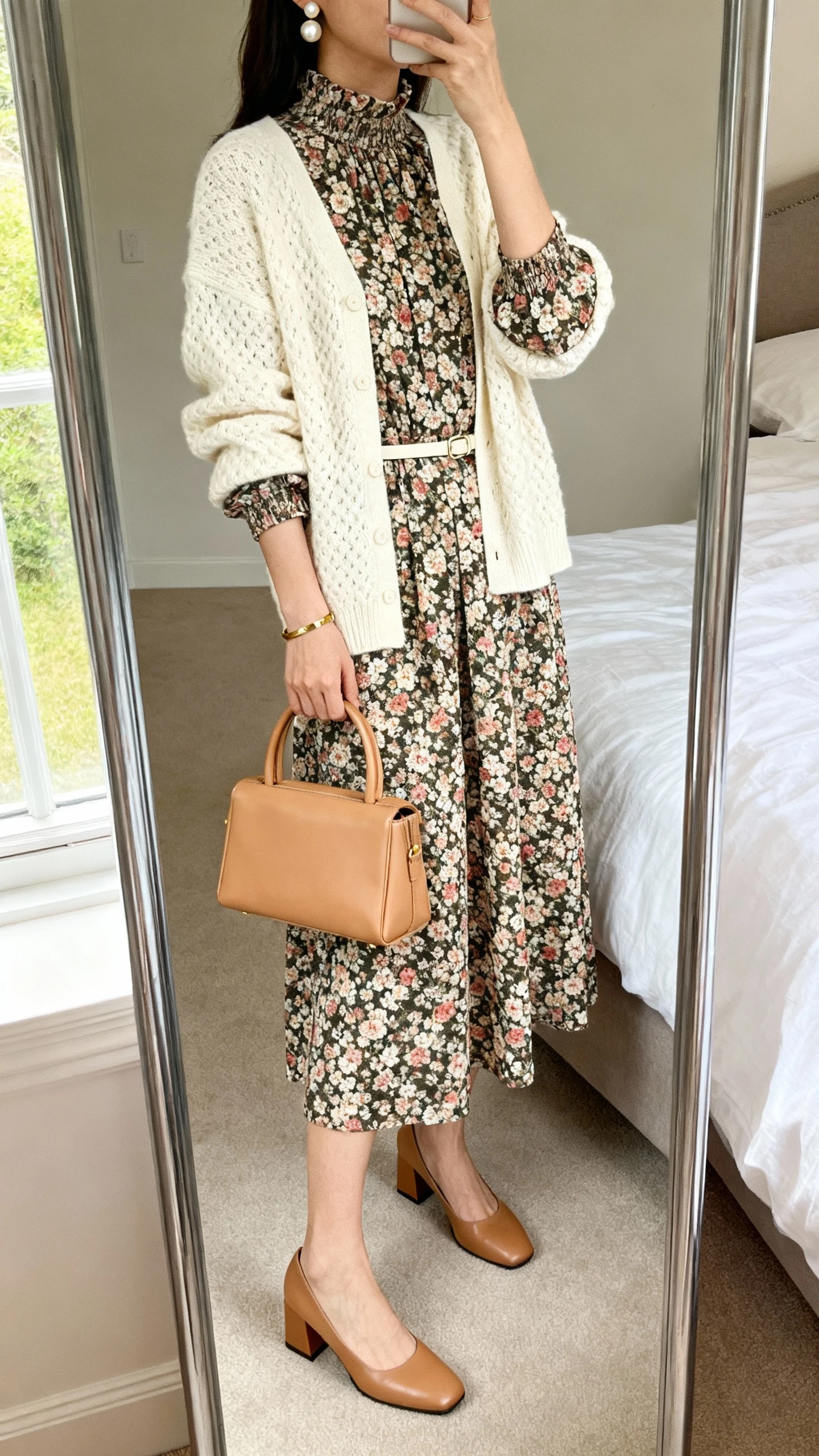iPhone mirror selfie of a woman wearing a floral midi dress with sleeves and a high neckline, a cream textured cardigan, block-heel Mary Janes, a structured top-handle bag in tan, pearl earrings, a delicate gold bracelet, and a thin waist belt, face not visible, cropped at neck, bedroom mirror, natural window light, iPhone photo quality.