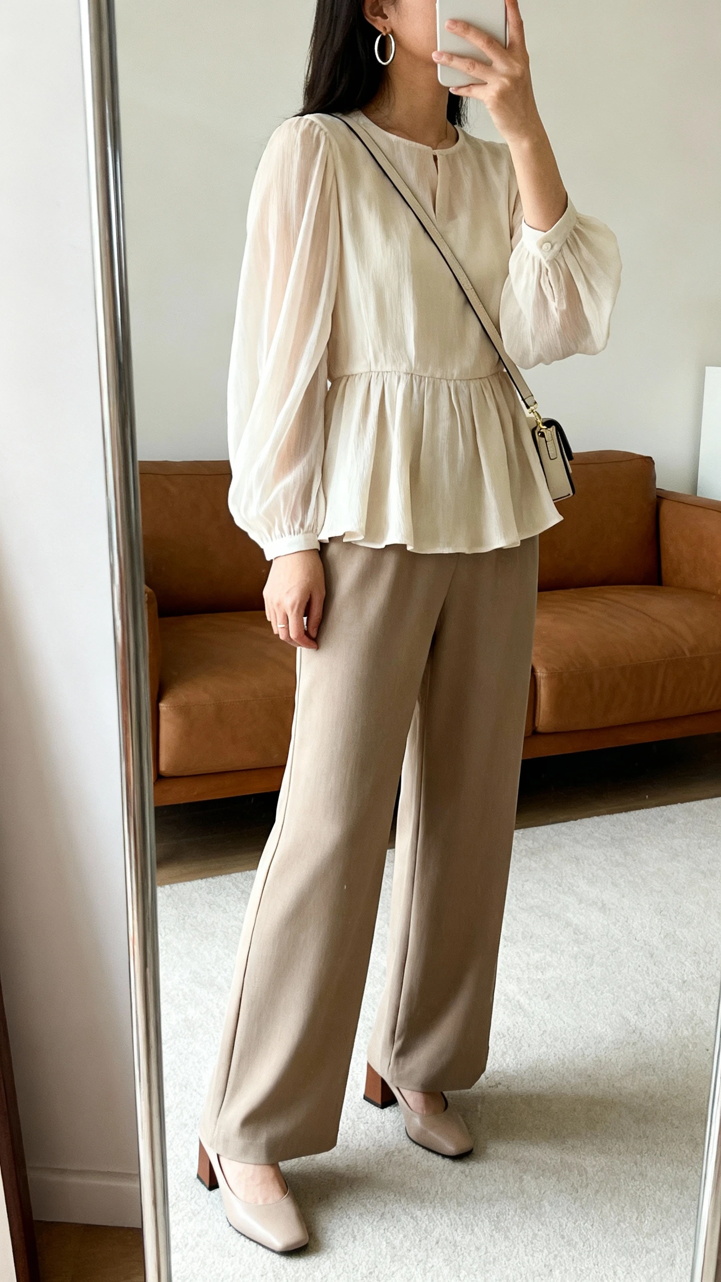 iPhone mirror selfie of a woman wearing a flowing peplum blouse with wide-leg trousers, slim crossbody bag, hoop earrings, and block heels, face not visible, side angle, living room full-length mirror, soft natural daylight, iPhone photo quality.