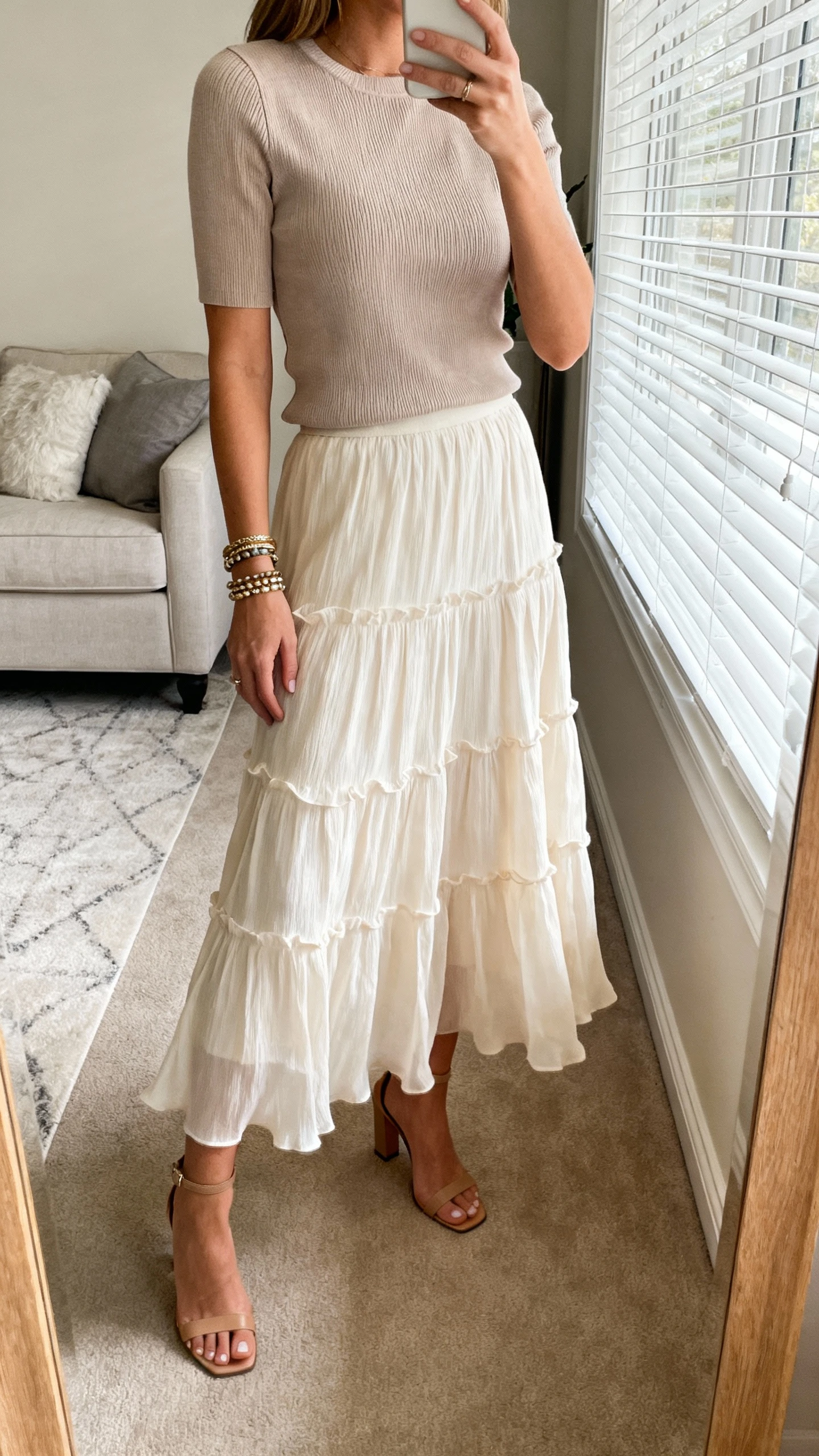 iPhone mirror selfie of a woman wearing a flowing tiered skirt with a structured top (tailored short-sleeve or fitted knit), heeled sandals, bracelet stack, face not visible, cropped at neck, living room mirror with soft window light, iPhone photo quality.